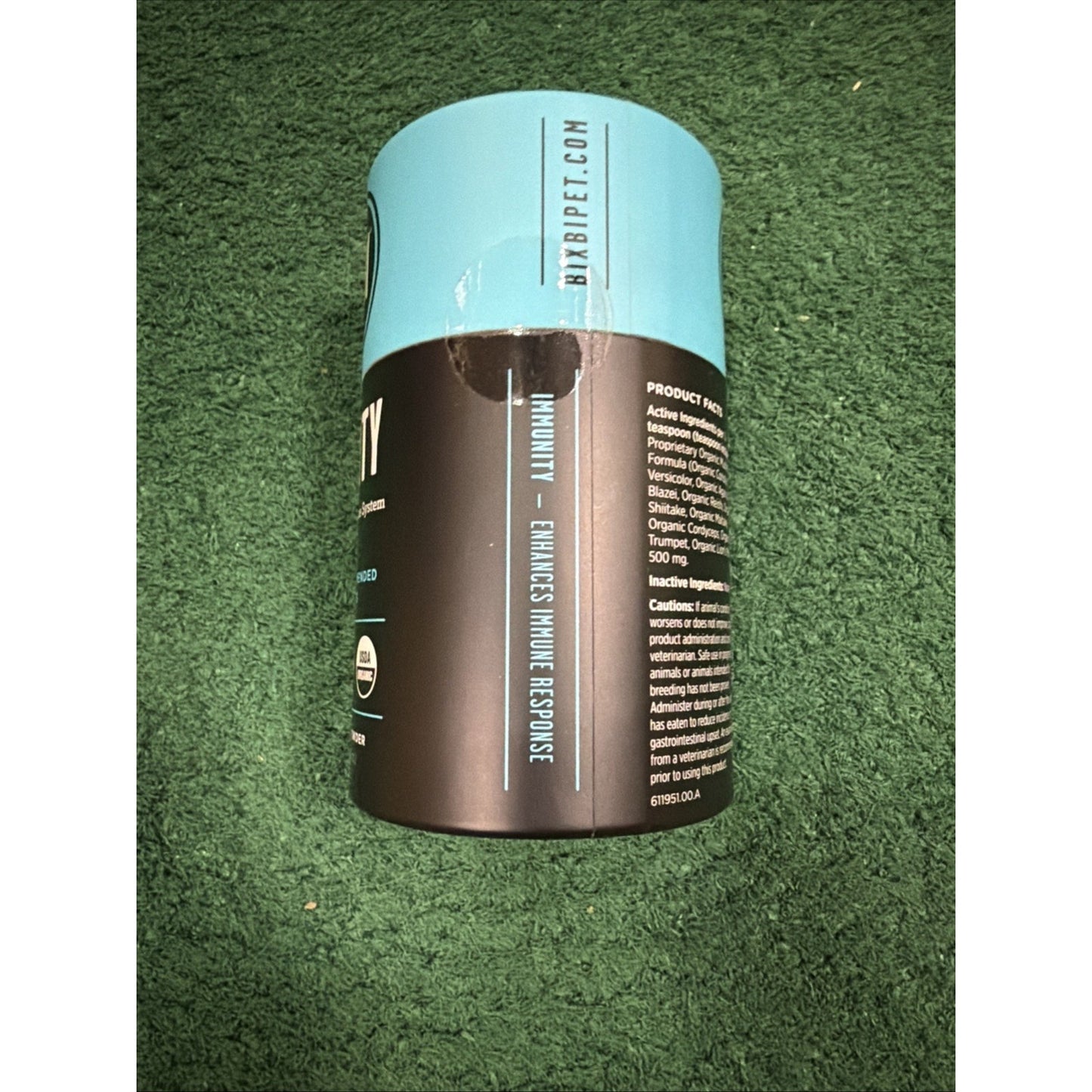 Cylindrical supplement container with a black and light blue label placed on a green textured surface, showing product information and branding details.