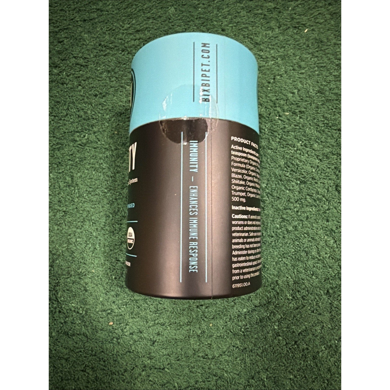 Cylindrical supplement container with a black and light blue label placed on a green textured surface, showing product information and branding details.