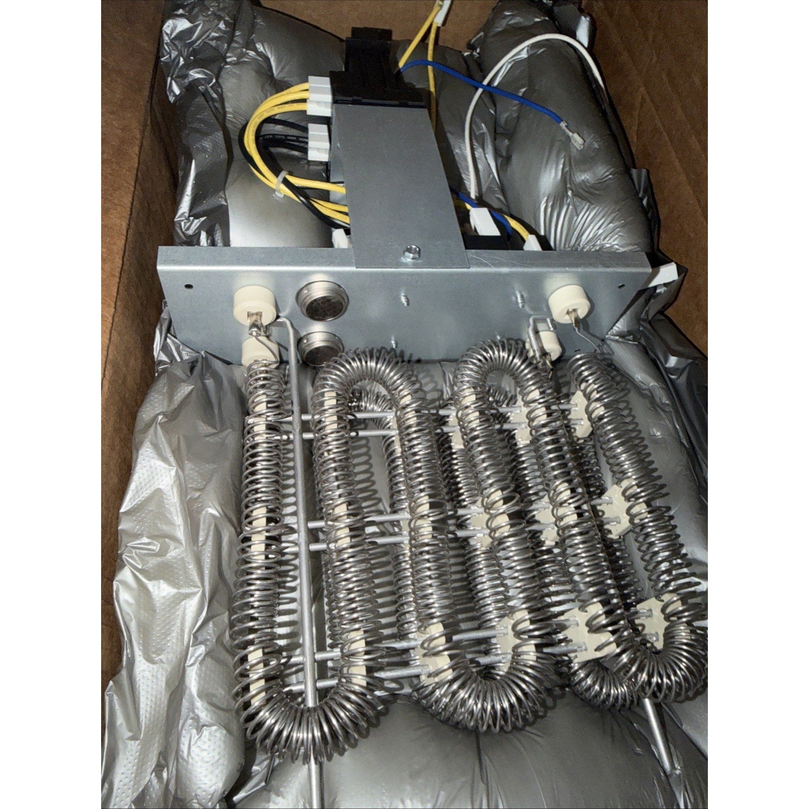 Metal electric heating coil assembly with multiple tightly wound coils mounted on a metal frame, connected to electrical wiring, resting inside a protective silver insulated packaging.