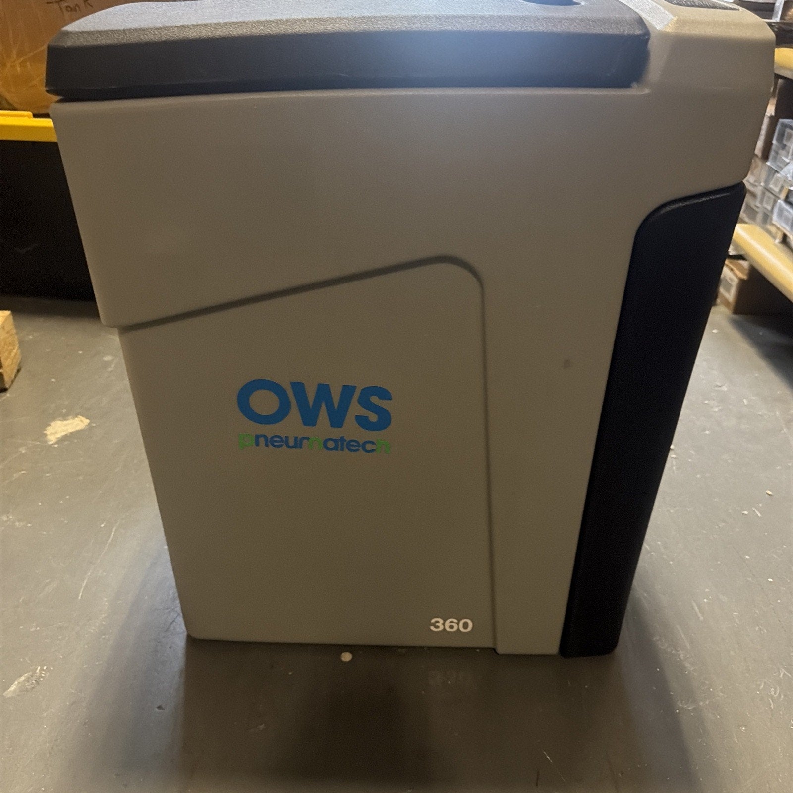 Gray and black industrial machine with the label "OWS pneurnatec 360" positioned on a workshop floor with shelving and tools in the background.
