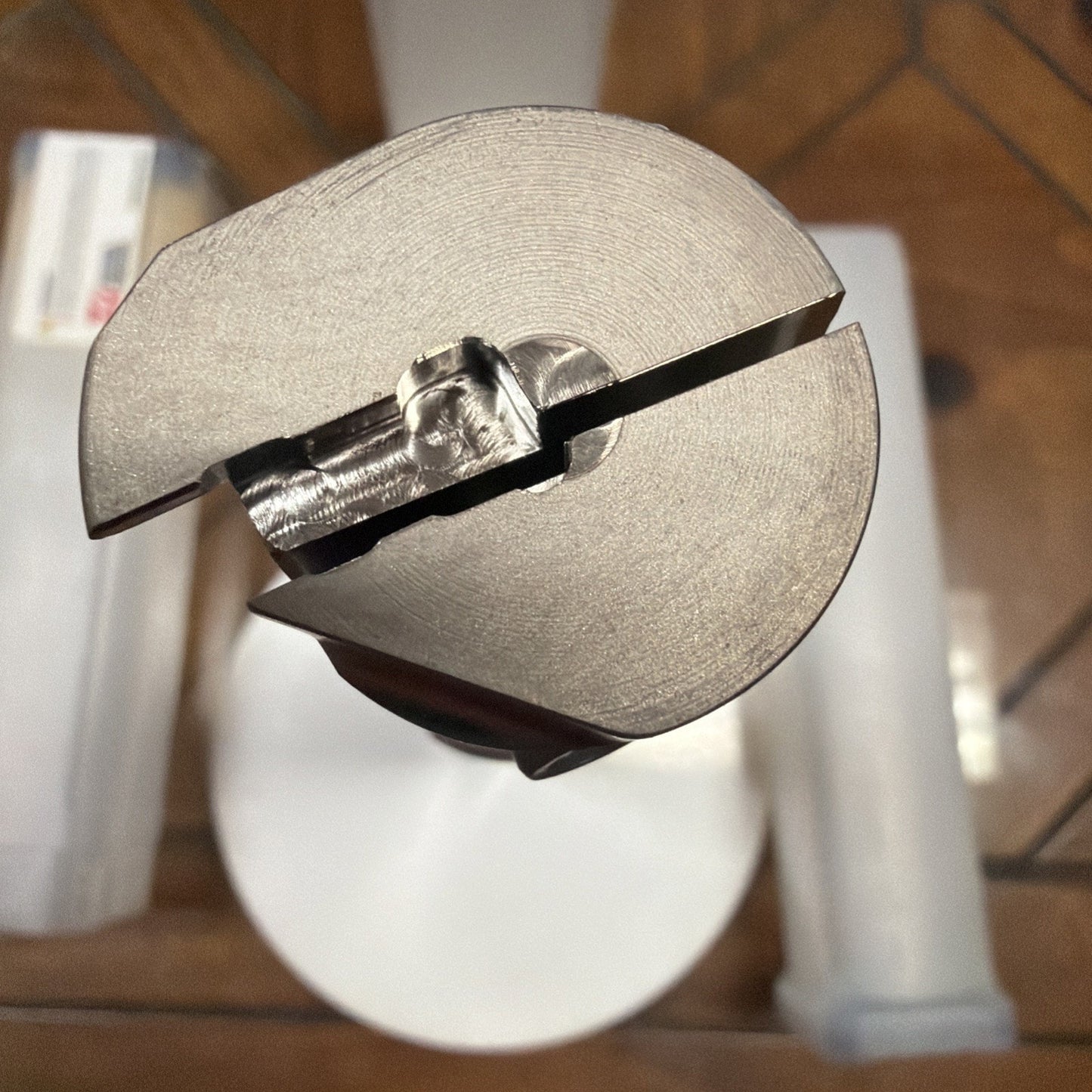 Close-up view of a cylindrical metal cutting or shaping tool with a precision-machined tip featuring a split and a reflective surface, placed on a wooden and glass table.