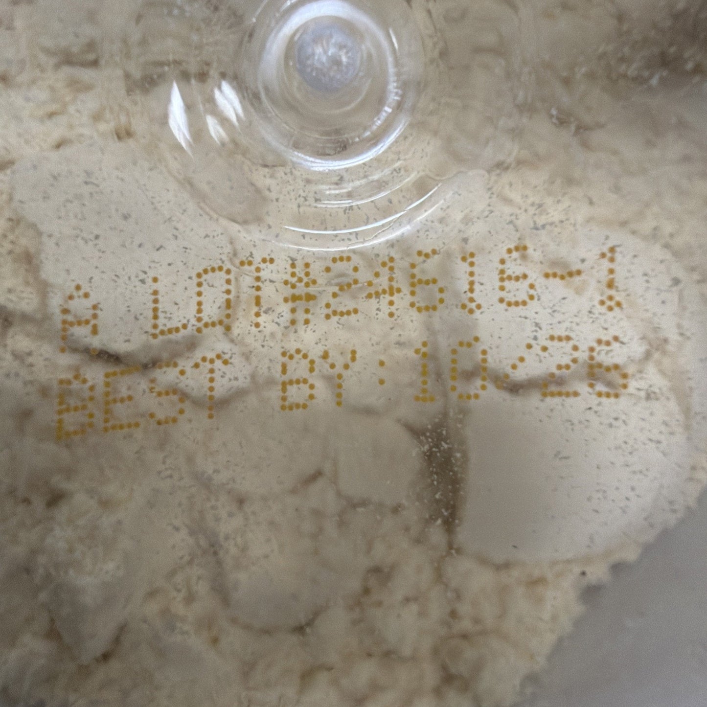Close-up of beige powder inside a clear container with a printed lot number and best-by date in yellow ink on the surface beneath the plastic lid.