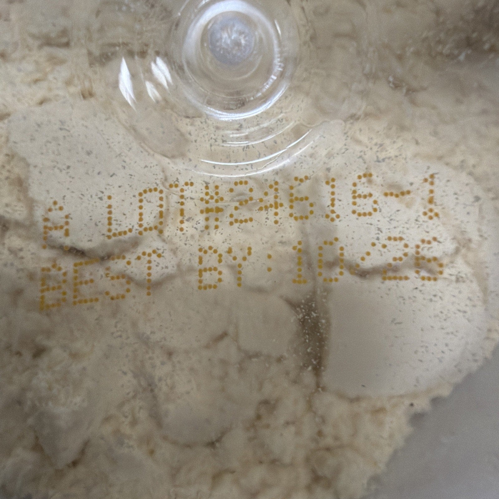 Close-up of beige powder inside a clear container with a printed lot number and best-by date in yellow ink on the surface beneath the plastic lid.
