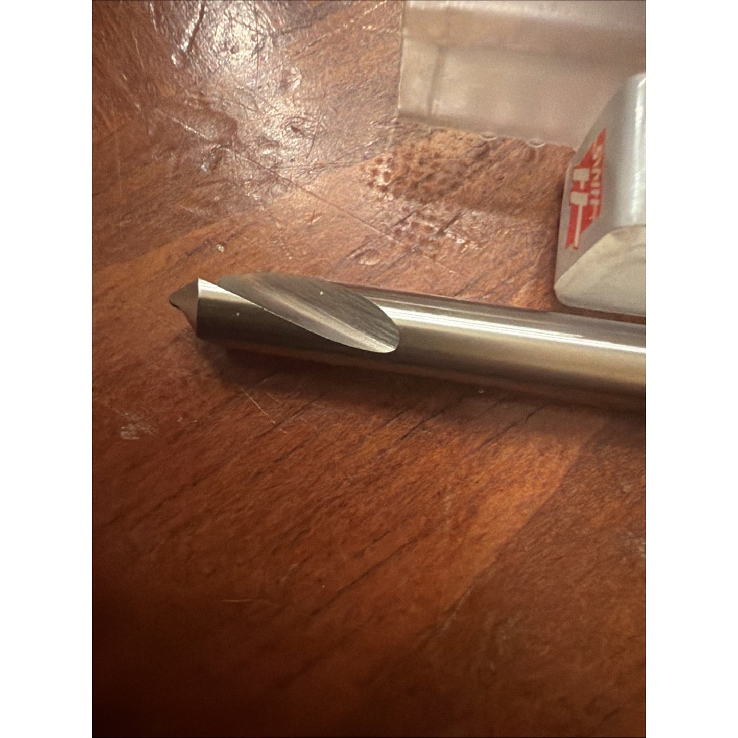 Close-up of a metal drill bit with a sharp pointed tip and spiral grooves, resting on a wooden surface next to a partially visible plastic case with a red and white label.