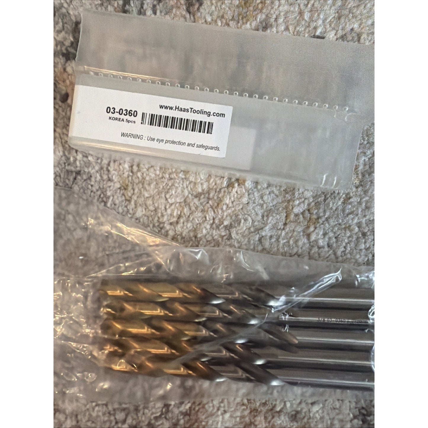 Five twist drill bits with gold-colored tips and silver shanks, packaged inside a clear plastic sleeve, resting on a textured light-colored fabric surface next to a labeled plastic bag.