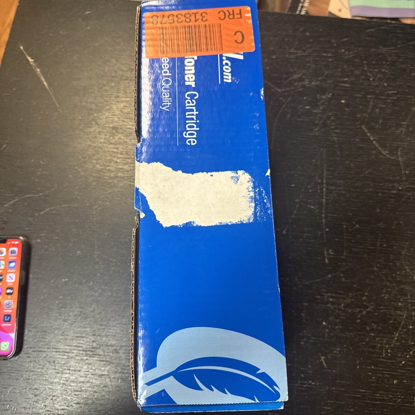 Blue rectangular box with partially visible white text reading "Toner Cartridge" and a feather graphic on the lower portion, placed on a dark surface with an orange barcode sticker on top.