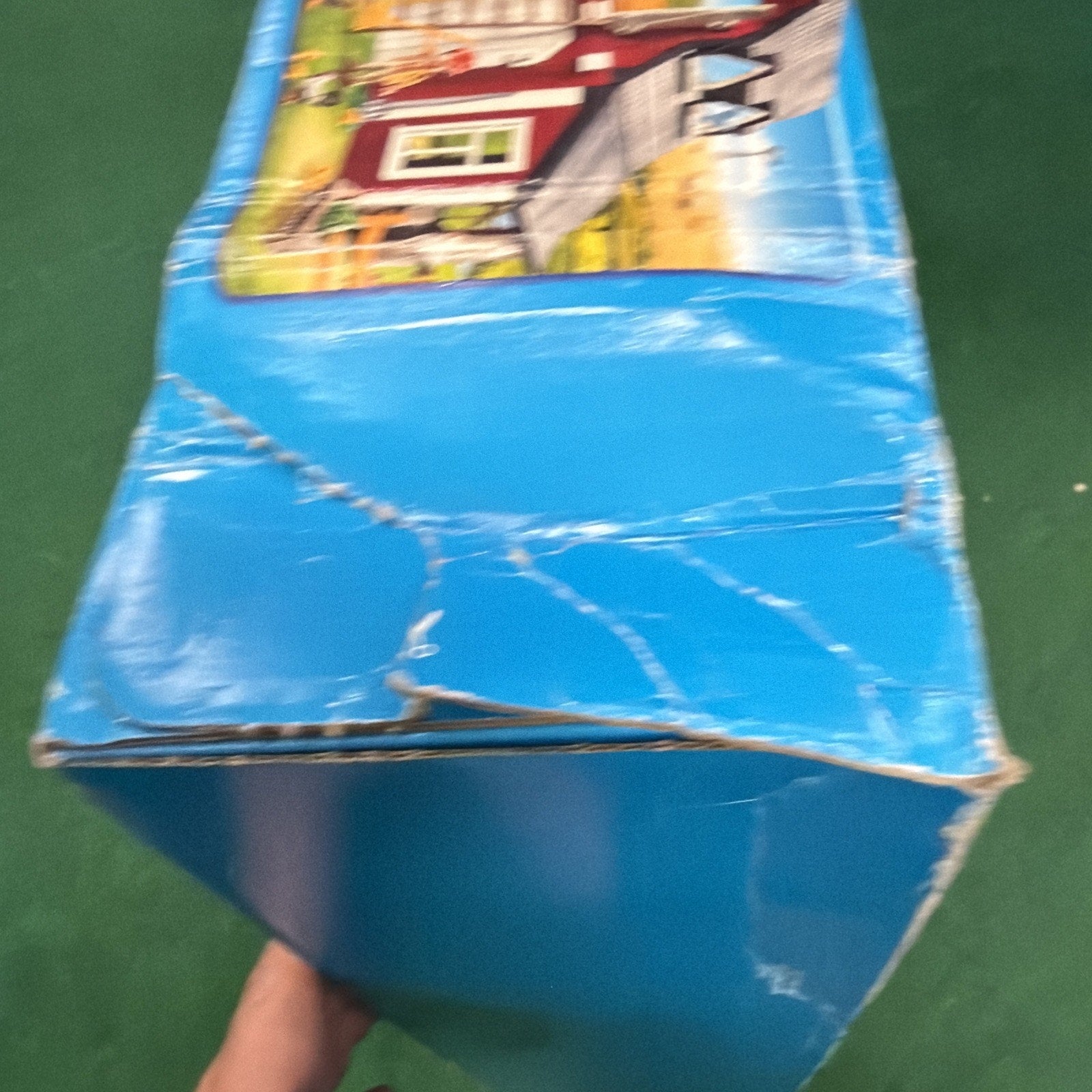 Close-up of a worn and creased blue cardboard box with visible edge wear, held by a hand against a green background, featuring part of a colorful printed design on one side.