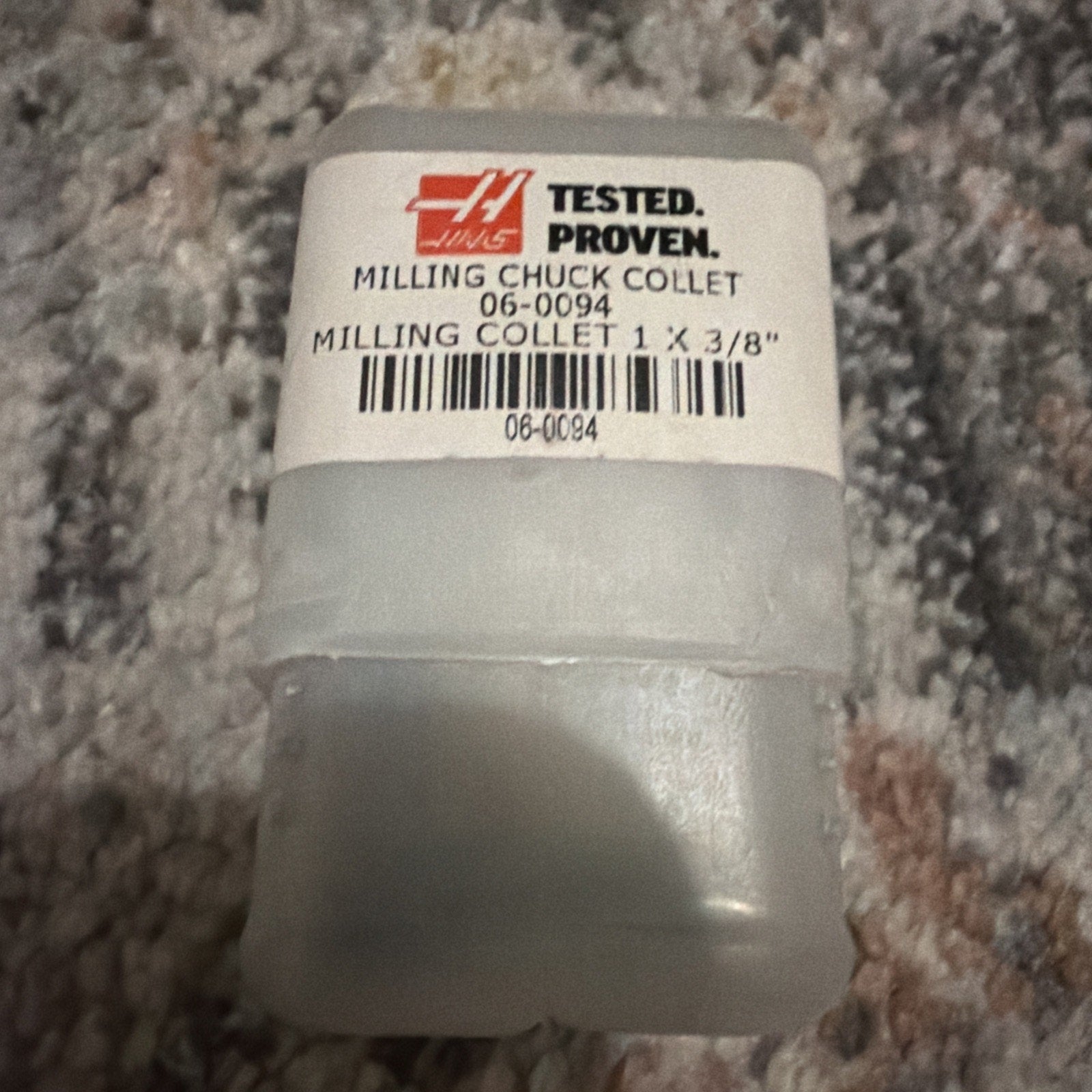 Opaque plastic container with a white label showing the Haas logo, product information for a milling chuck collet size 1 x 3/8 inch, and a barcode, resting on a textured carpet surface.