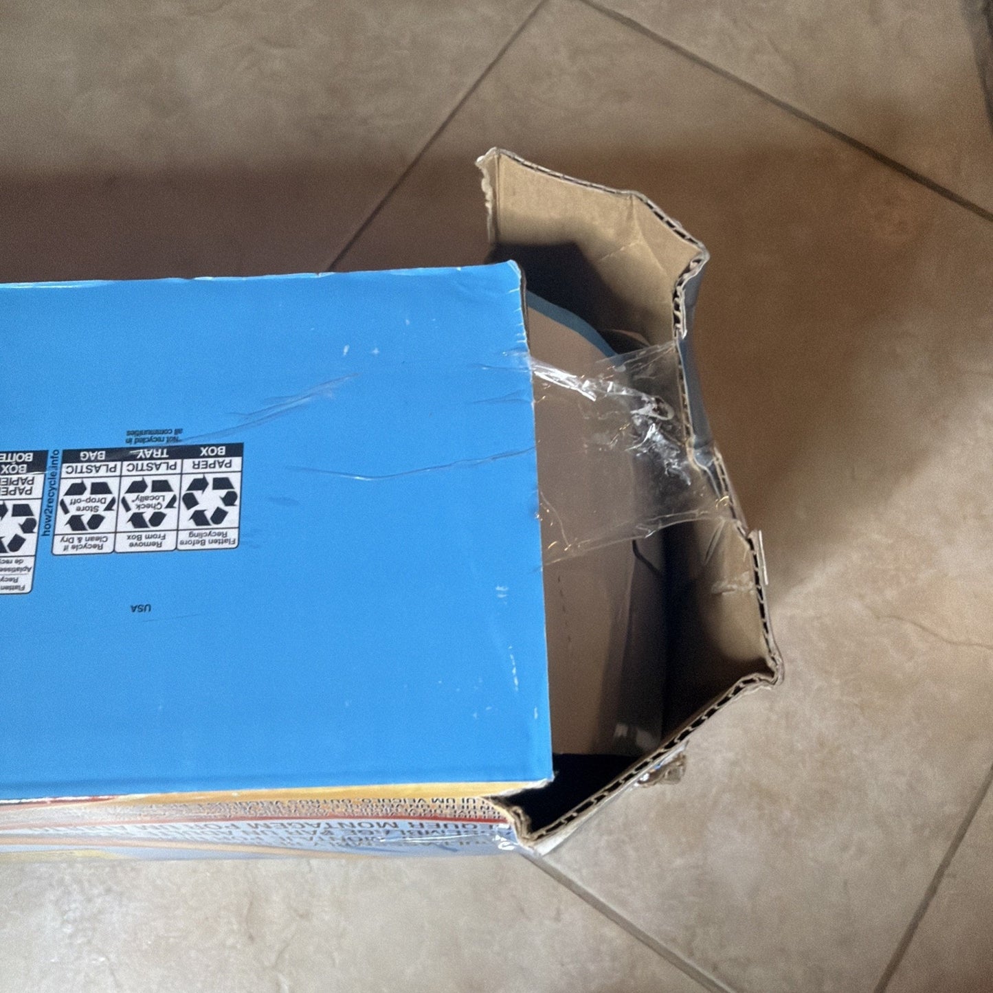 Partially opened brown cardboard shipping box with clear packing tape sealing an edge, resting on a tiled floor with visible recycling symbols on the blue inner flap.