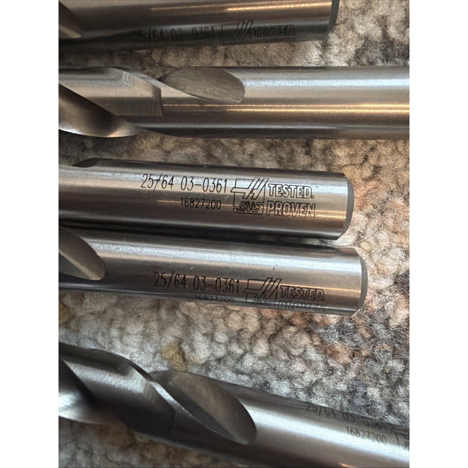 Close-up of several silver metal drill bits with engraved size markings and "TESTED" labels, lying on a textured beige carpeted surface.