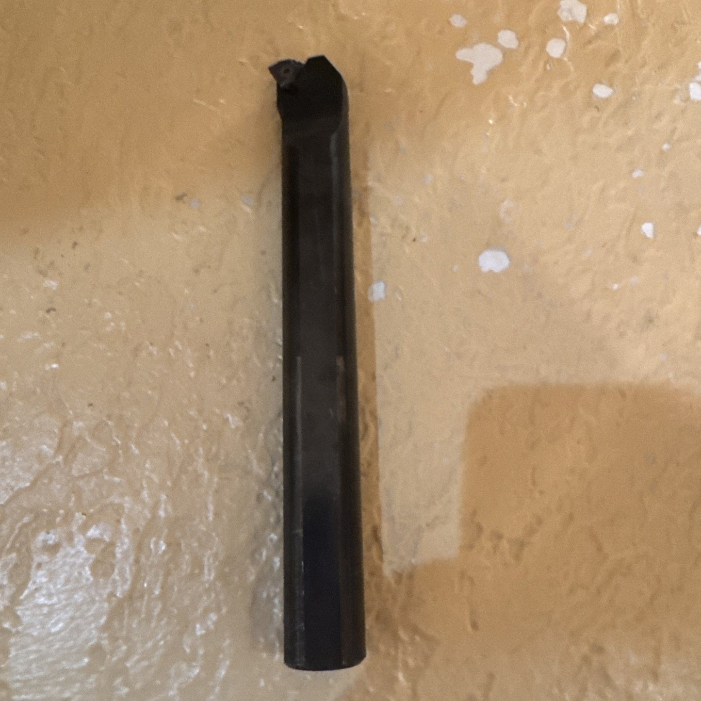 Single dark metal cutting tool with a worn and chipped tip, standing vertically against a textured beige surface with scattered white spots.