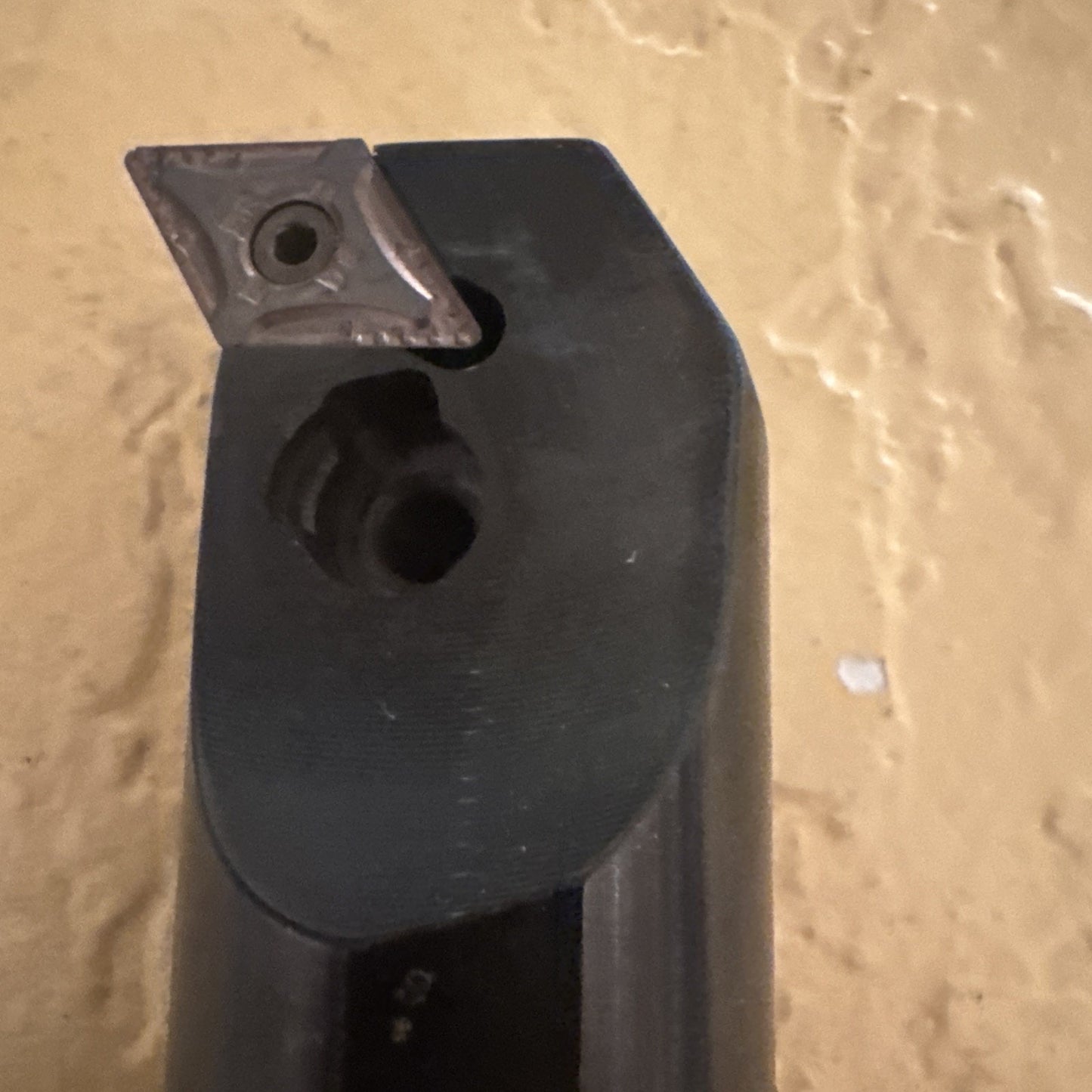 Close-up of a black metal cutting tool with a square carbide insert secured by a screw, positioned against a textured beige wall background.