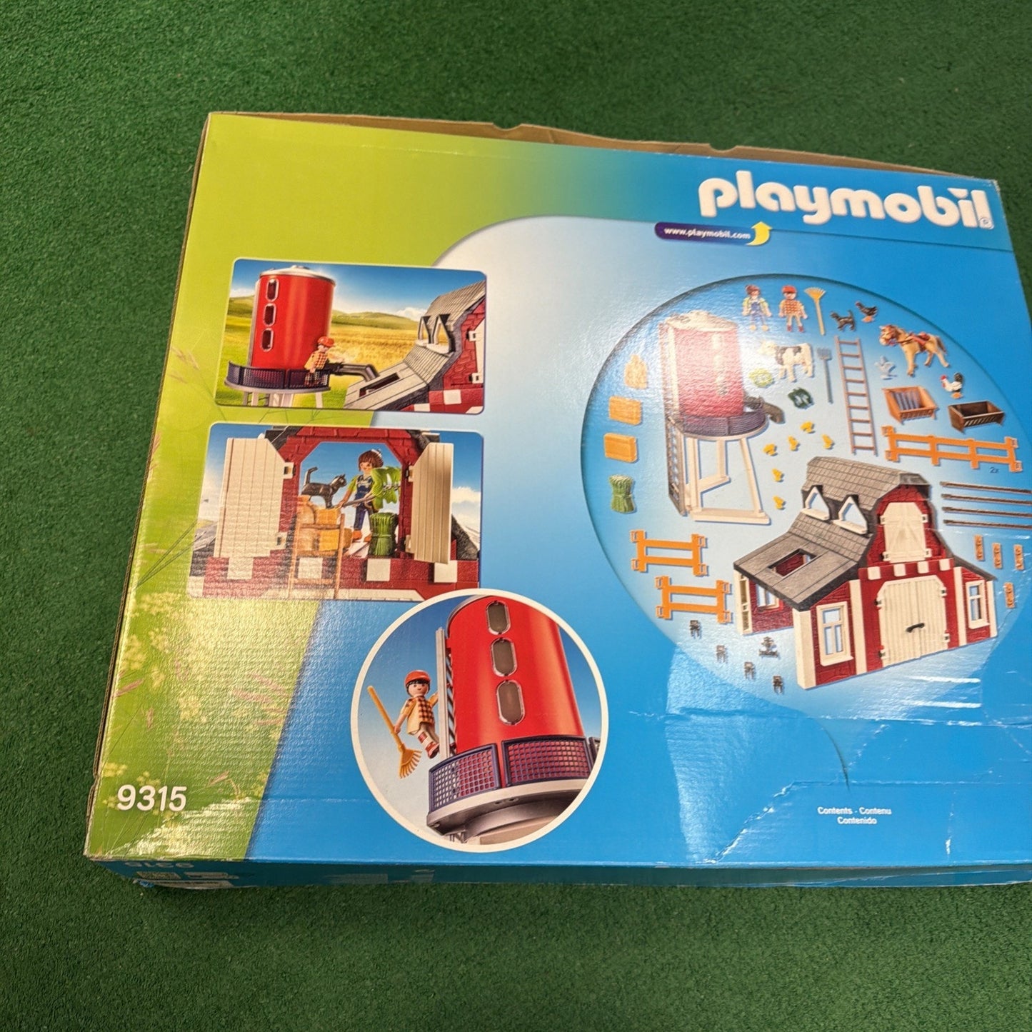 Box of Playmobil toy set featuring a red cylindrical silo, a small barn with white doors, farm animal figures, fences, ladders, and multiple accessories, displayed on a blue and green background.