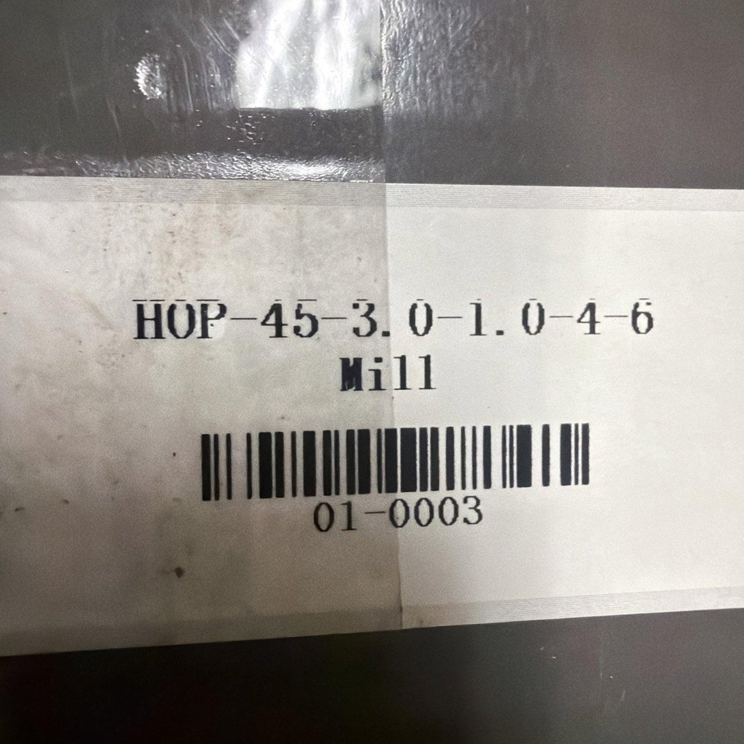 Close-up of a white label with black printed text and barcode reading "HOP-45-3.0-1.0-4-6 Mill" and "01-0003" on a dark plastic or metal surface.