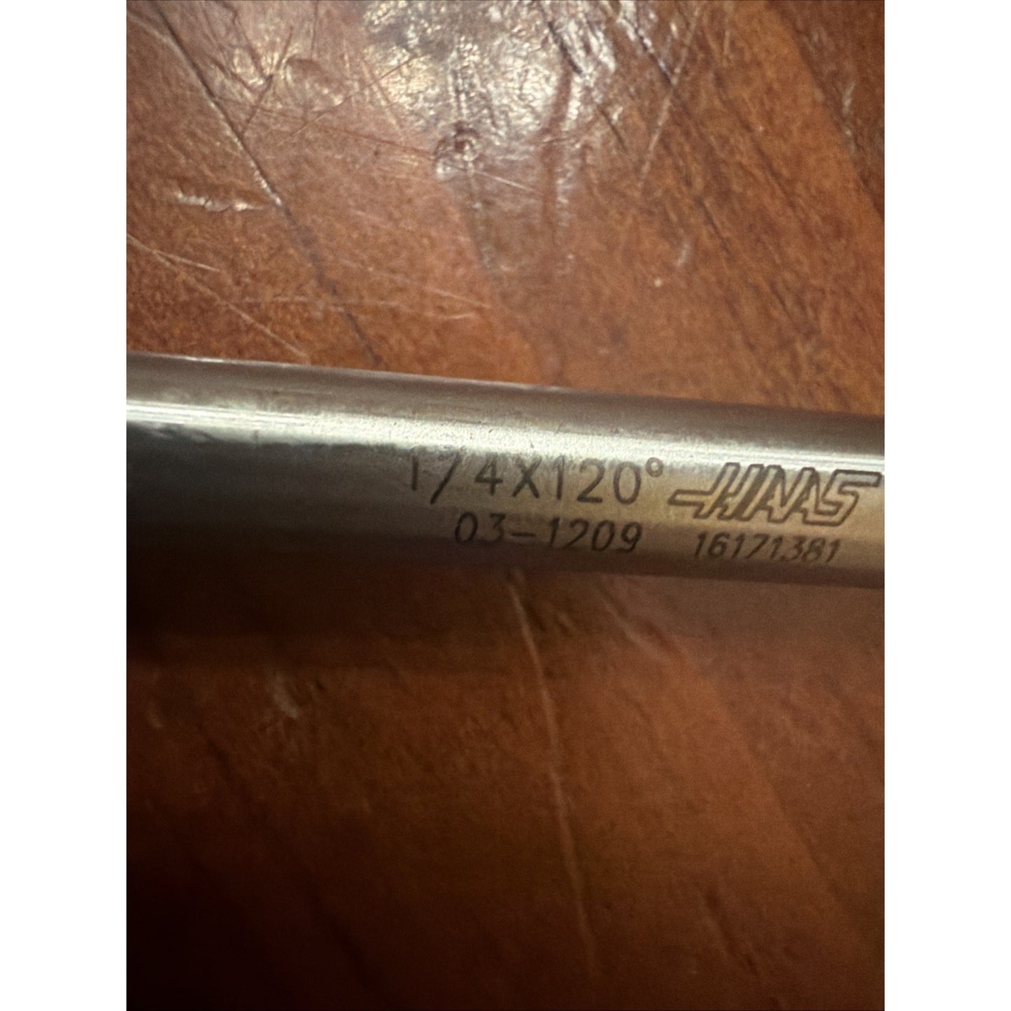 Close-up of a metal rod with engraved text "1/4X120°," a product code, and a logo, resting on a scratched brown surface.