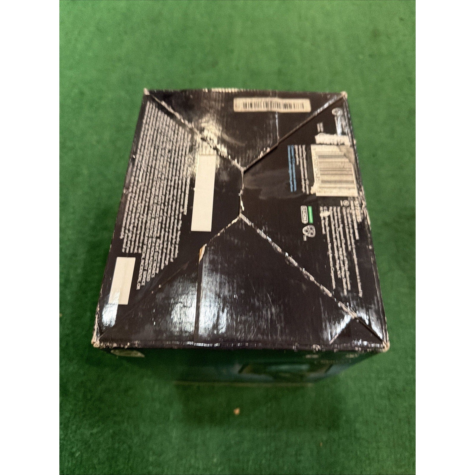 Dark-colored cardboard box with visible creases, dents, and wear on the top surface, placed on a green textured background.