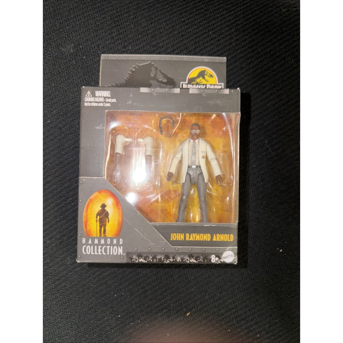 Action figure of John Raymond Arnold from Jurassic Park Hammond Collection, dressed in a white lab coat and suit, displayed in a black and orange collector’s box with Jurassic Park logo.