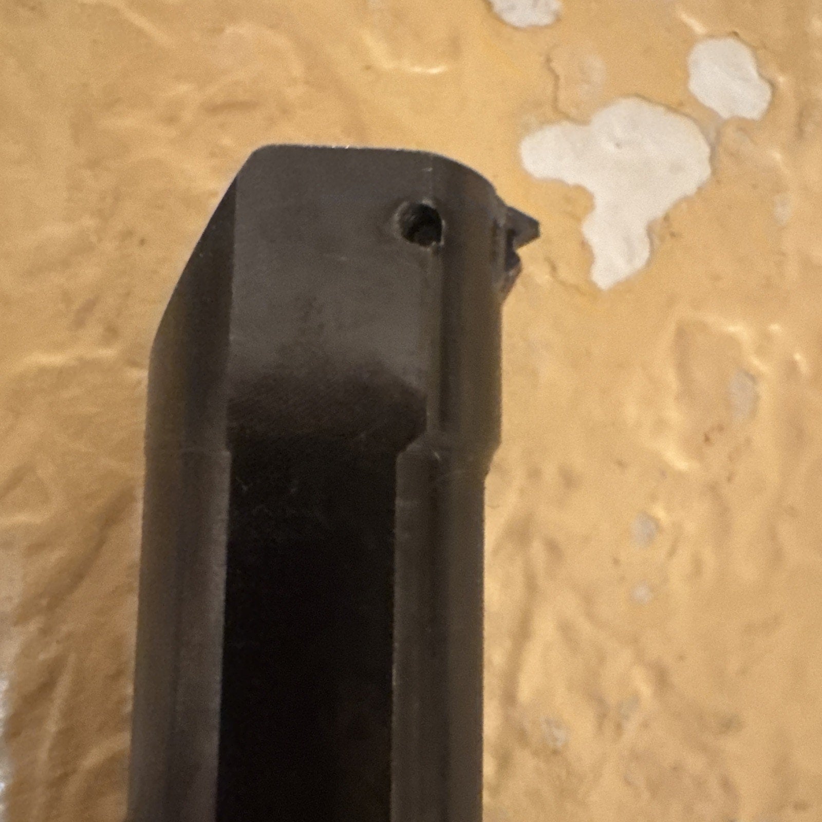 Close-up of a black metal cutting tool with a small, angled cutting tip and a circular mounting hole near the edge, positioned vertically against a textured yellow-beige wall.