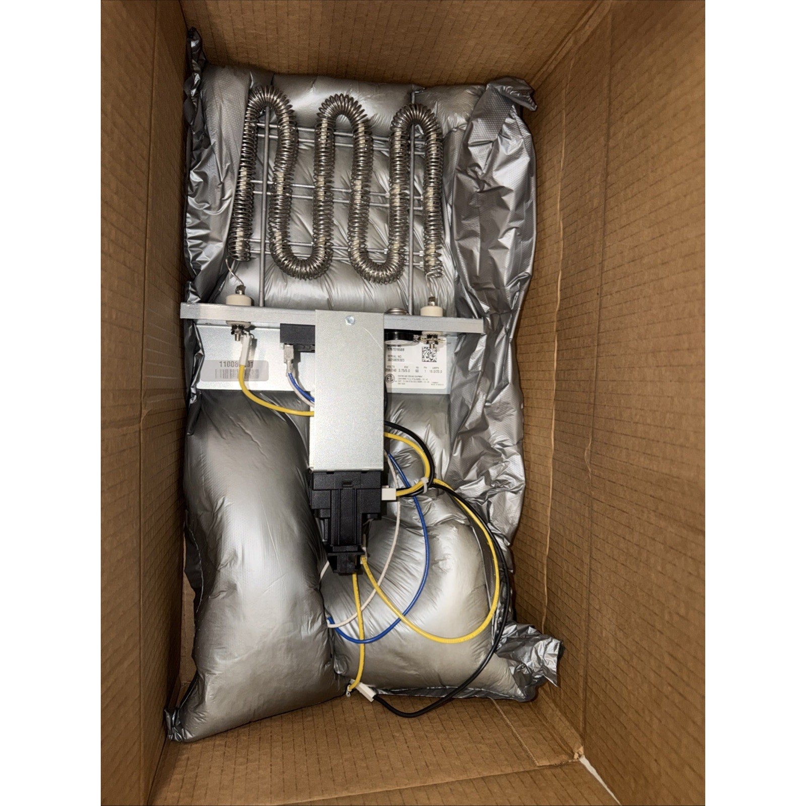 Electric heating element with coiled metal wire, mounted on a metal bracket with attached wiring, placed inside a brown cardboard box cushioned with silver protective packaging.