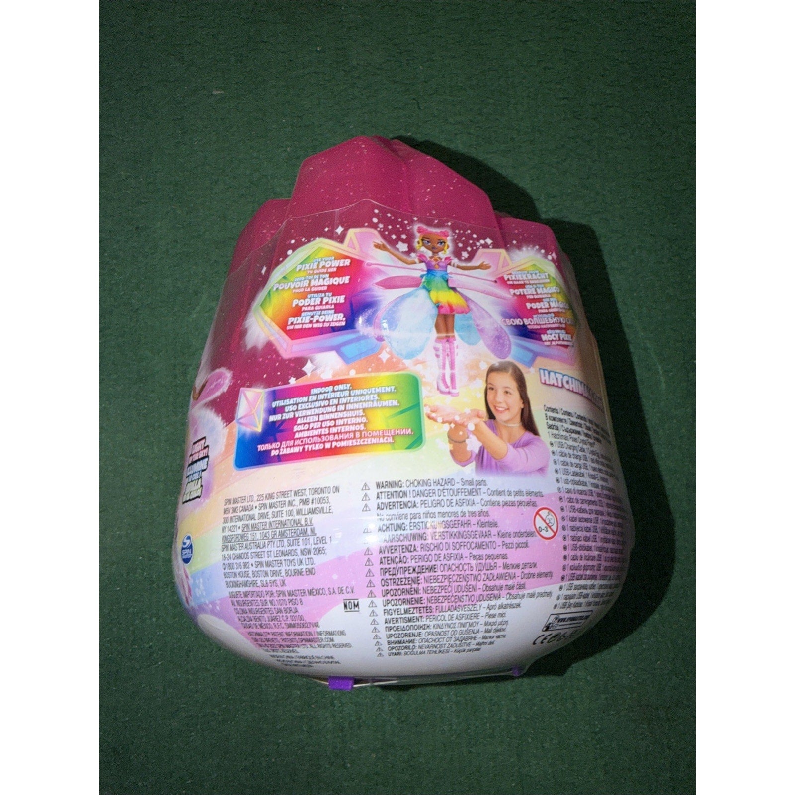 Pink and white toy packaging shaped like an egg, featuring a colorful fairy character illustration, product information, and a photo of a smiling girl holding the toy against a green background.