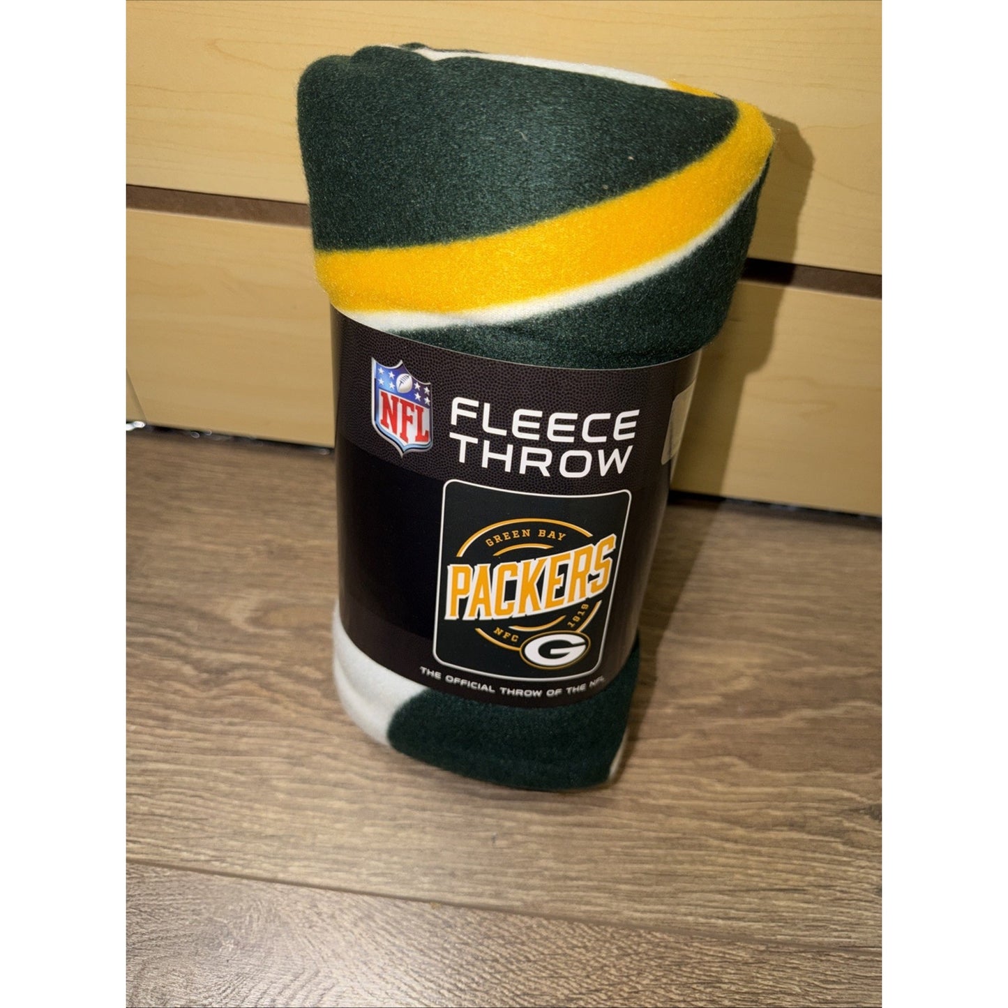 Rolled Green Bay Packers fleece throw blanket featuring green, yellow, and white stripes, wrapped with a black NFL-branded label on a wooden surface.