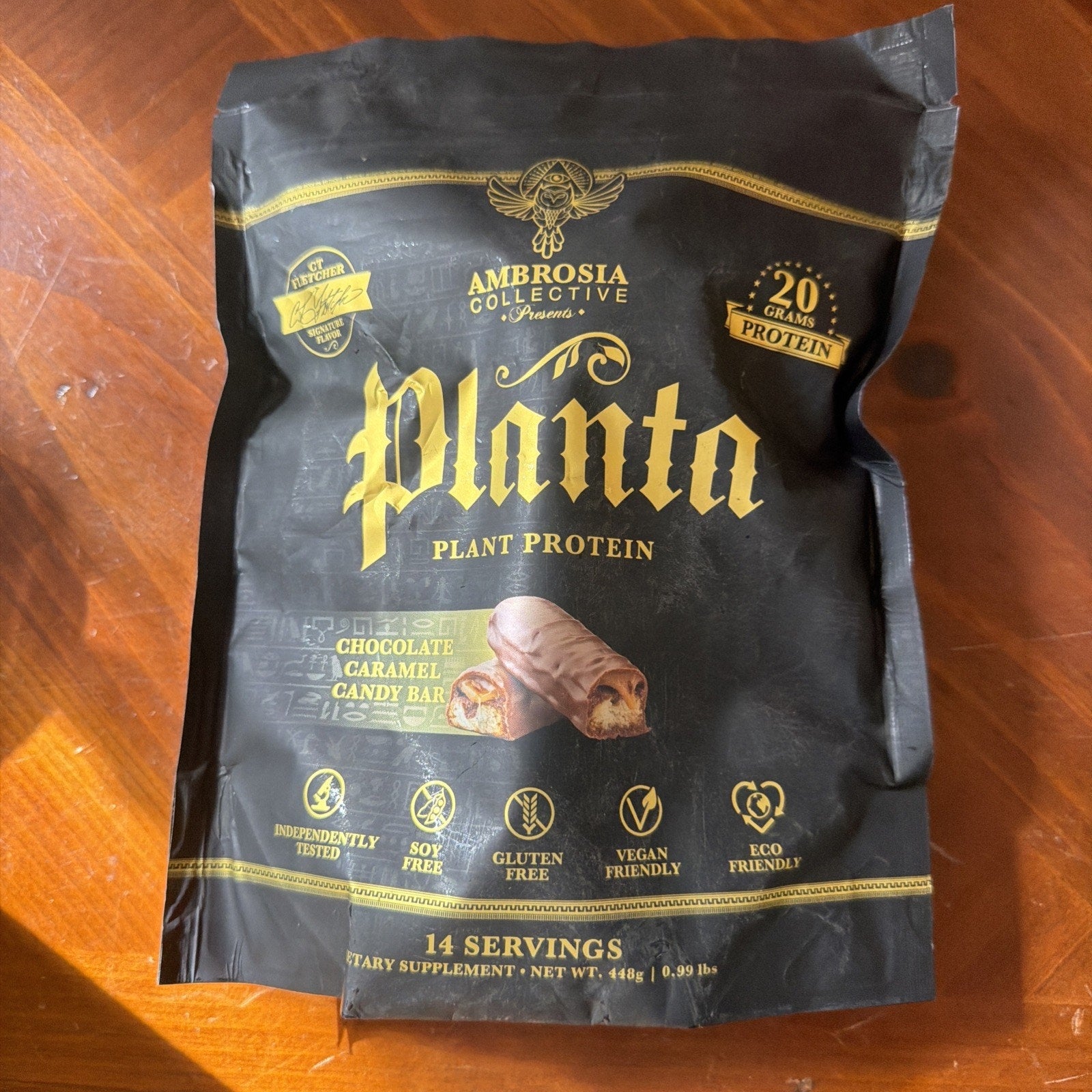 Black resealable bag of Ambrosia Collective Planta plant protein in chocolate caramel candy bar flavor, featuring 20 grams of protein per serving and icons for soy-free, gluten-free, vegan, and eco-friendly attributes.