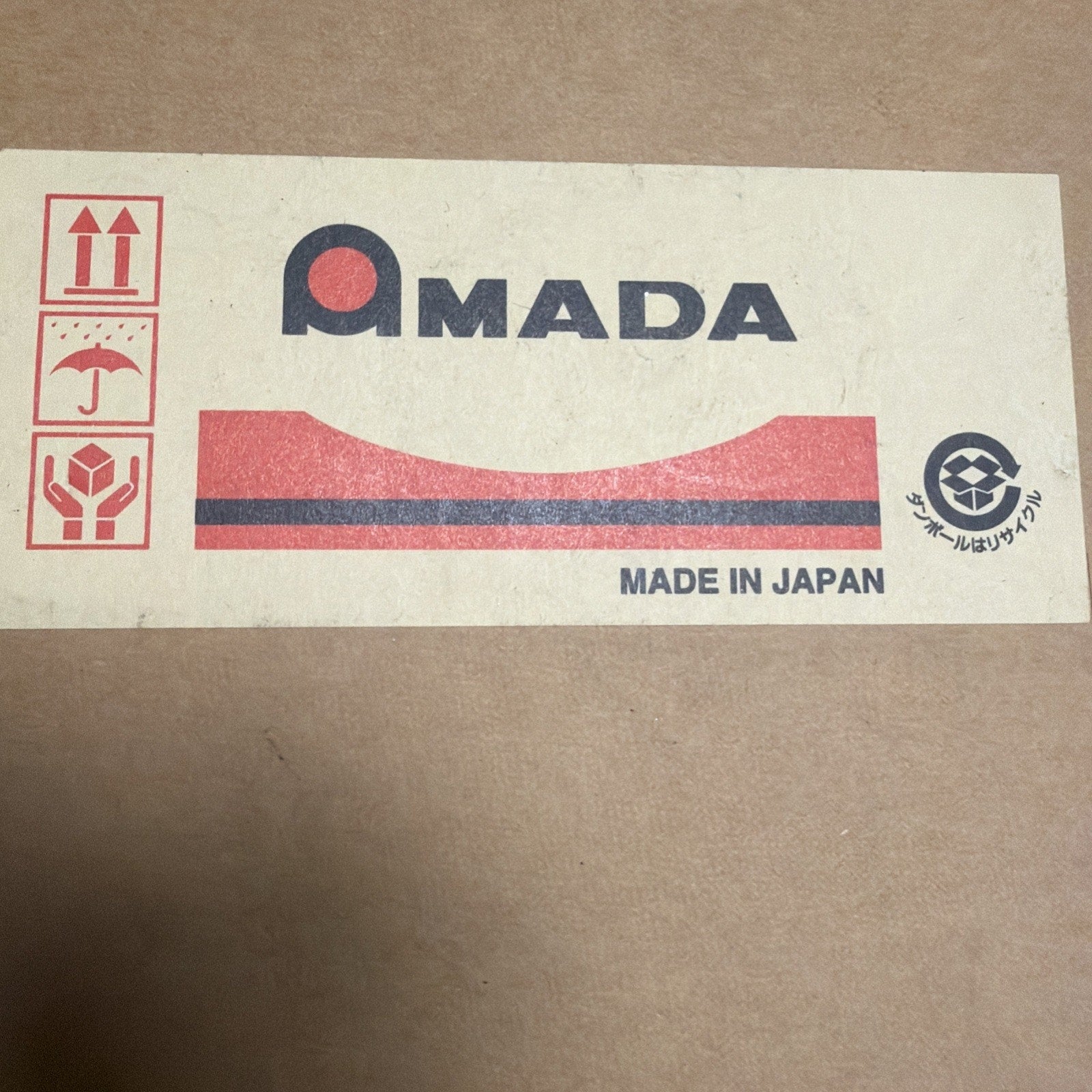 Beige shipping label on a cardboard surface featuring red handling icons for upright, keep dry, and fragile, with blue and red "AMADA" logo and "Made in Japan" text.