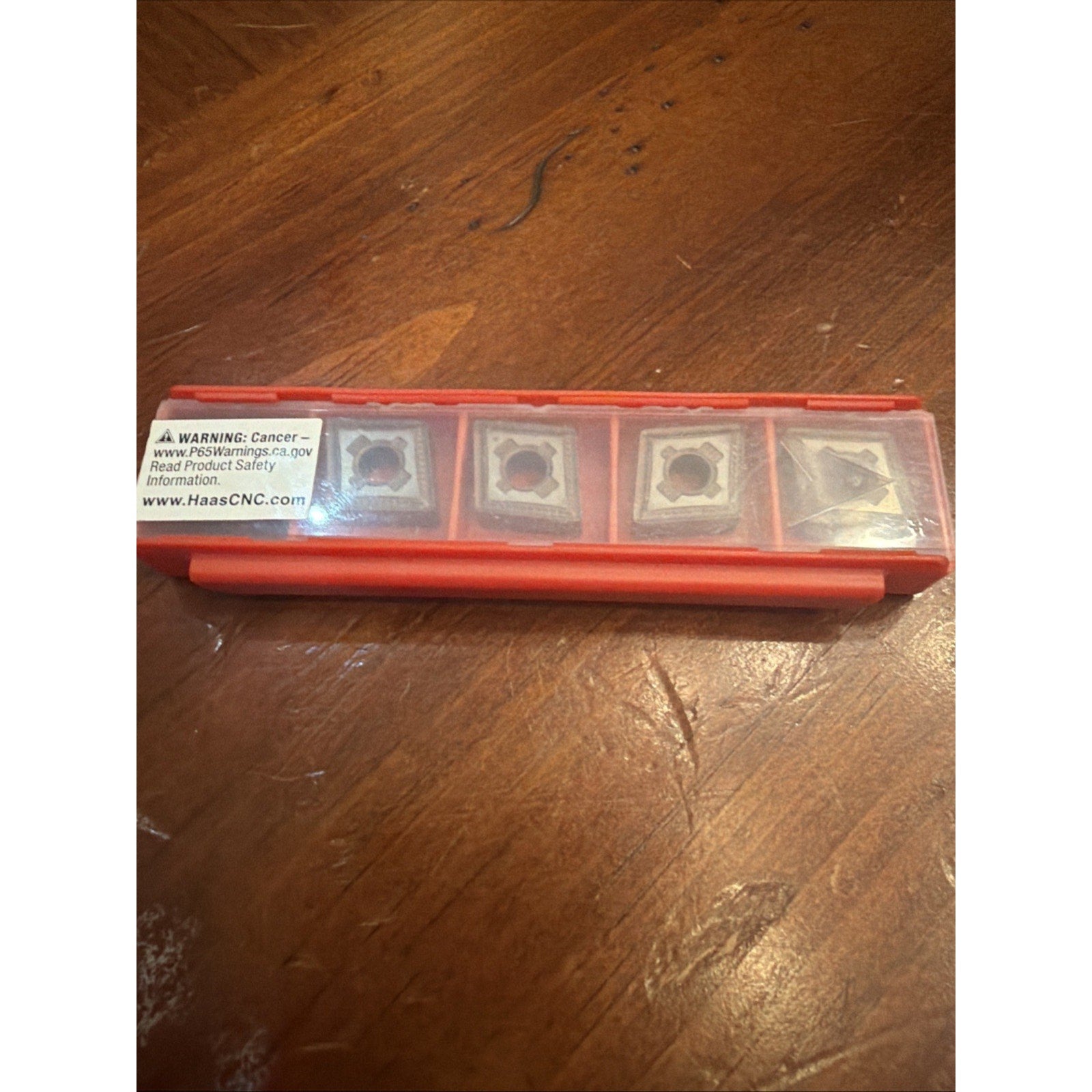 Red plastic case with a transparent lid containing five square metal cutting inserts, each with a central hole and visible wear, resting on a wooden surface.