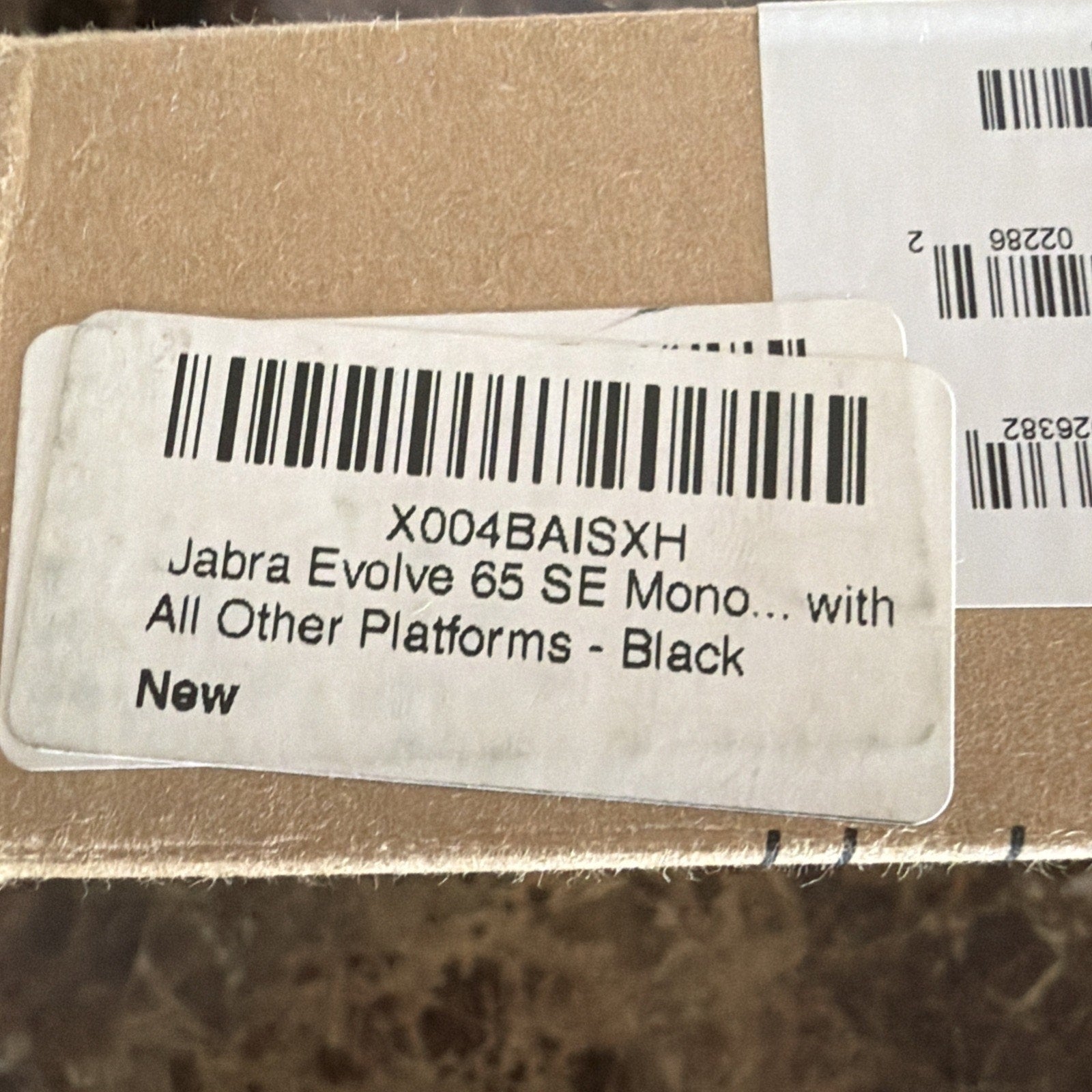 Close-up of a white product label on a brown cardboard box displaying a barcode, model code, and partial text indicating a black mono headset compatible with multiple platforms, marked as new.