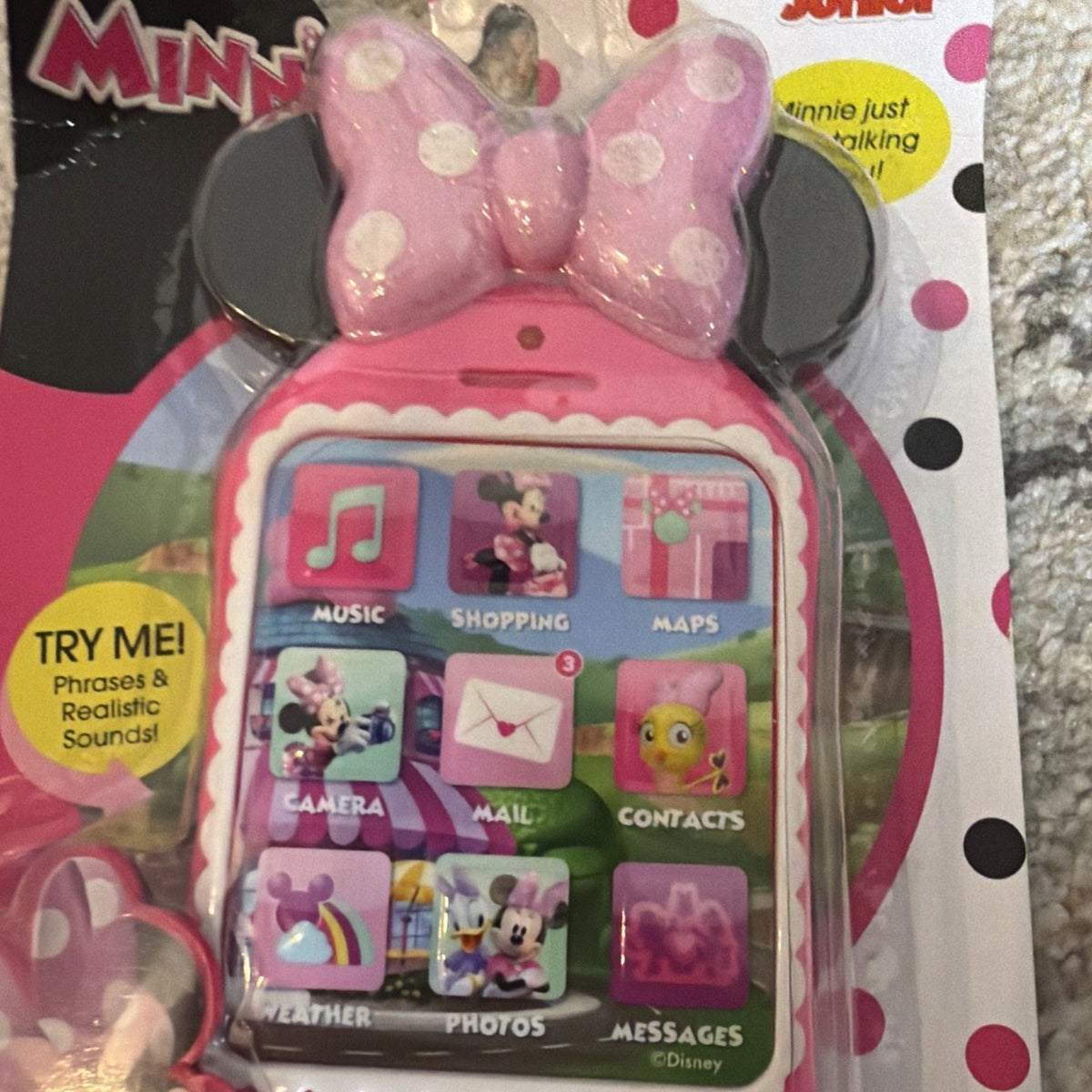 Child’s toy phone shaped like Minnie Mouse’s head with large pink polka dot bow, featuring colorful icons for music, shopping, maps, camera, mail, contacts, weather, photos, and messages, packaged with a try-me button for phrases and sounds.