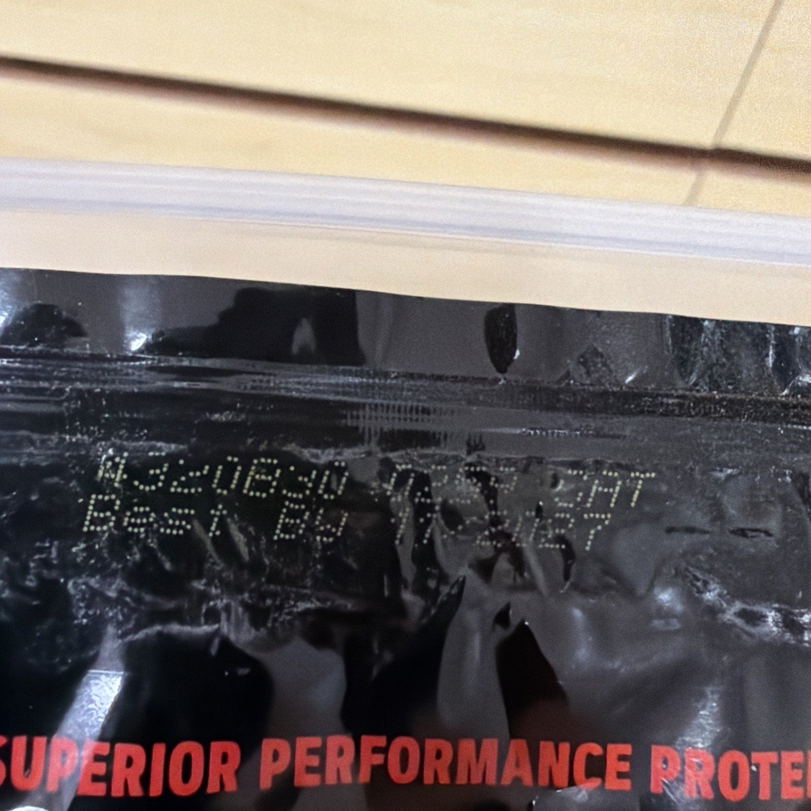 Close-up of black packaging with a printed expiration date of August 20, 2024, and a best by date of December 3, 2027, above red text partially reading "SUPERIOR PERFORMANCE PROTEIN.