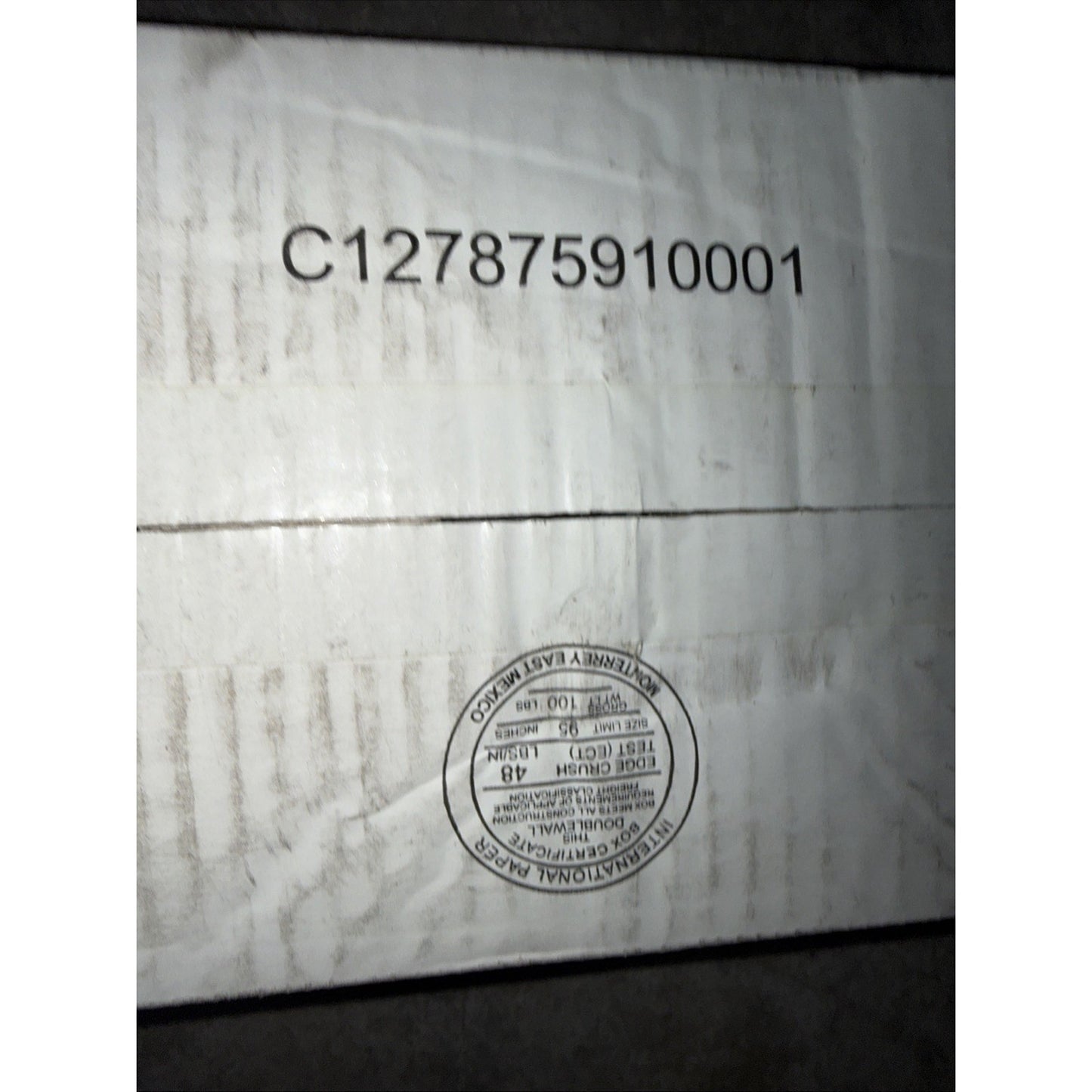 Close-up of a white cardboard box with a black printed code and a circular international packaging certification label indicating compliance and origin details.