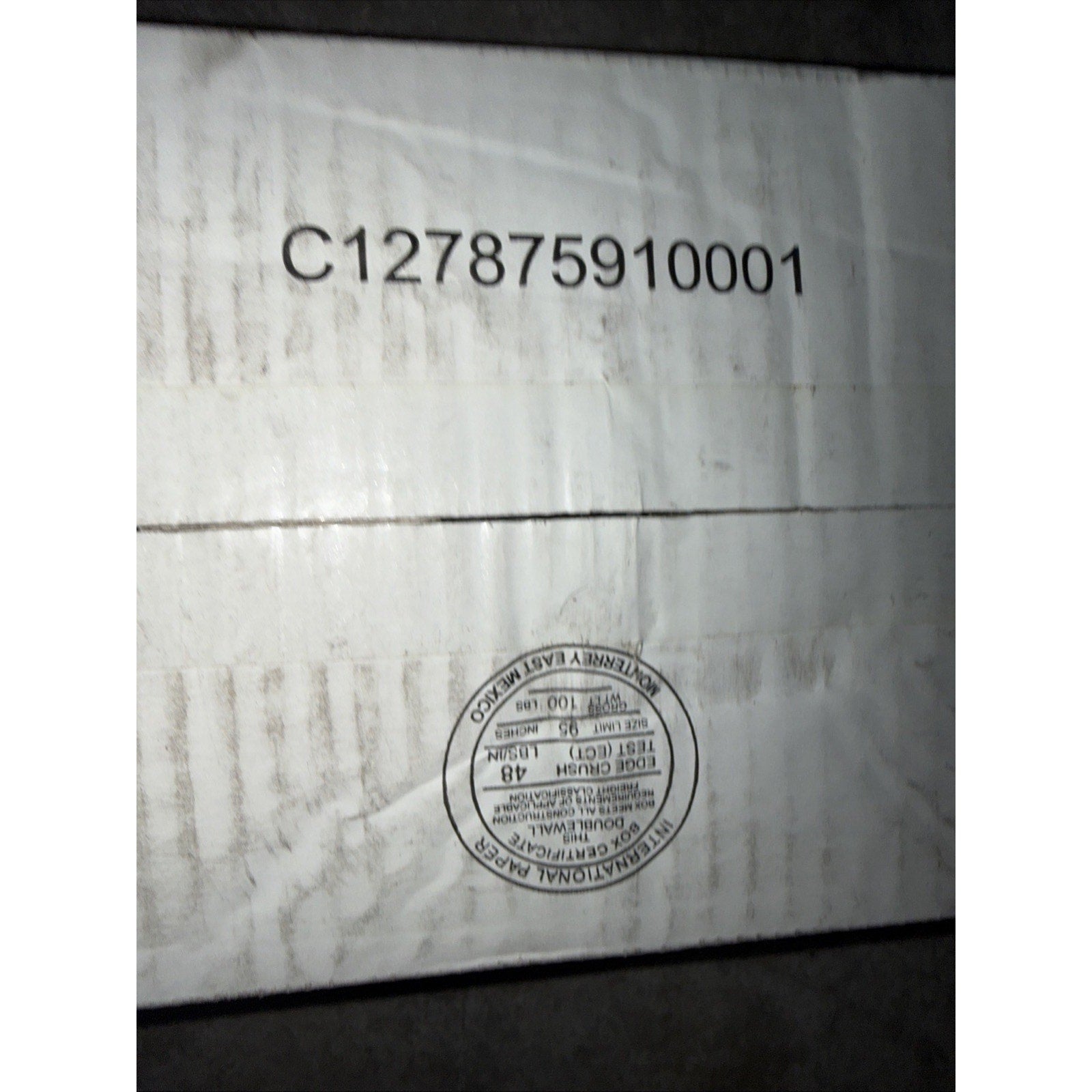 Close-up of a white cardboard box with a black printed code and a circular international packaging certification label indicating compliance and origin details.