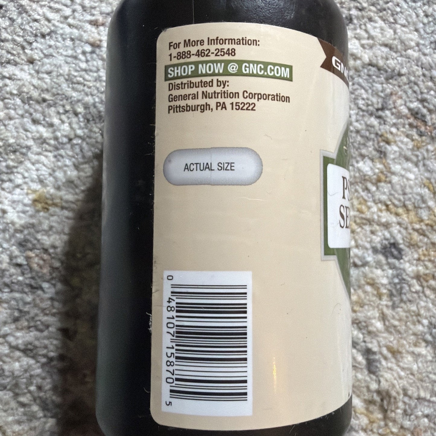 Close-up of a supplement bottle label showing contact information for General Nutrition Corporation, a website link to GNC.com, a barcode, and a pill displayed in a small window labeled "actual size.