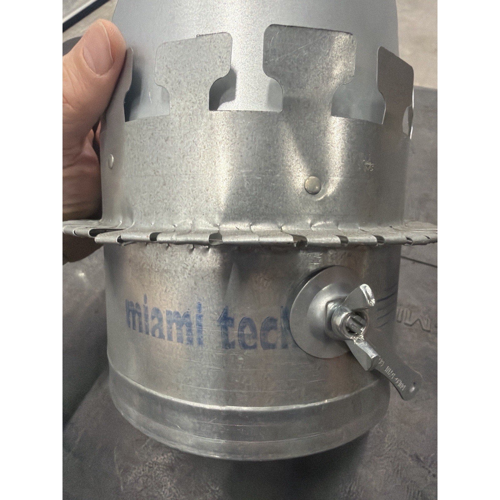 Close-up of a galvanized metal duct component featuring a circular clamp and a small wing nut valve handle, with faint blue text partially reading "miami tech" stamped on the surface.