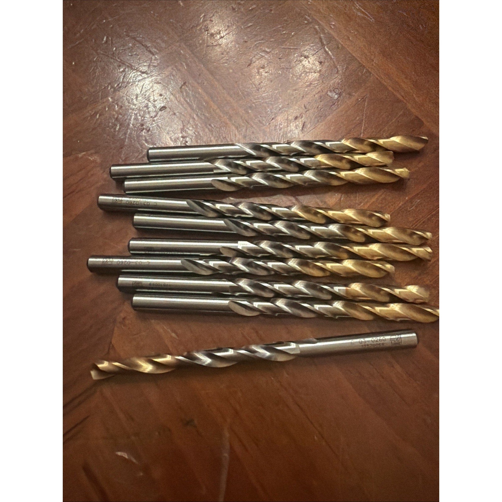 Ten metal drill bits with spiraled flutes, some coated in gold, arranged on a textured wooden surface, with one drill bit lying separately below the group.
