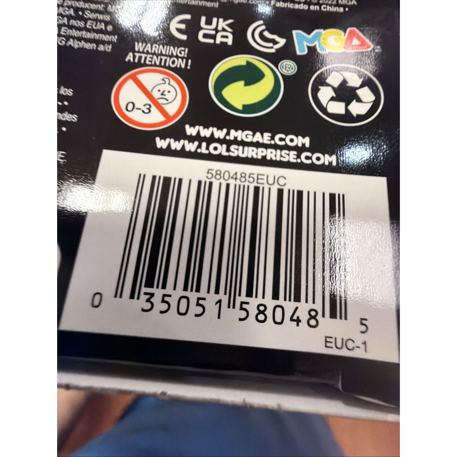 Close-up of a product barcode label with recycling and age warning symbols, website URLs, and brand logos printed on black packaging material.