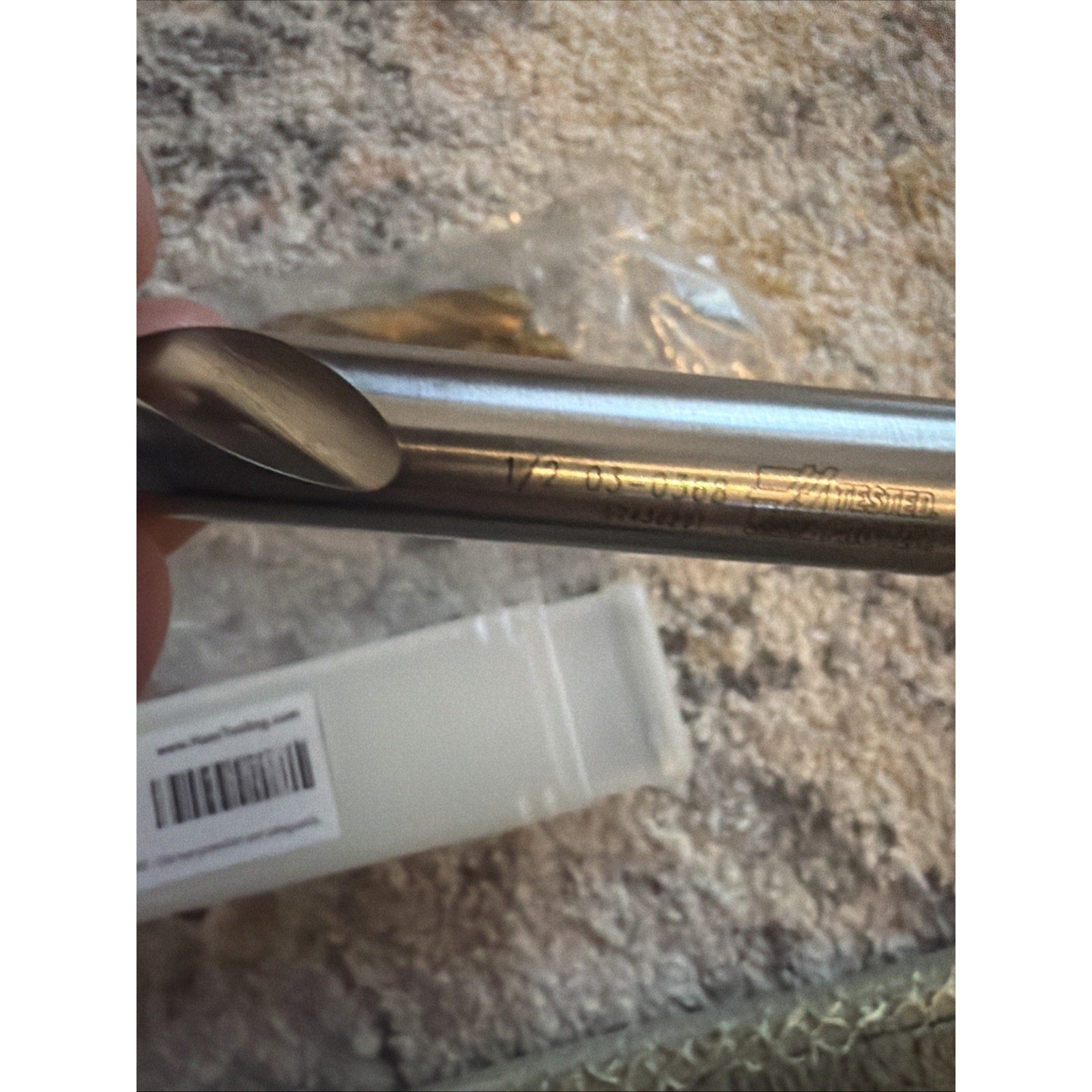 Close-up of a smooth silver metal rod with engraved text and numbers, held against a textured fabric background with a partially visible white plastic case and clear packaging nearby.