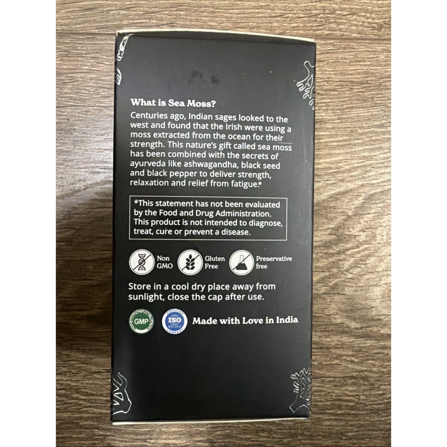 Back of a black sea moss dietary supplement box featuring product description, FDA disclaimer, icons for Non-GMO, Gluten-Free, Preservative-Free, storage instructions, and certification seals against a wooden background.