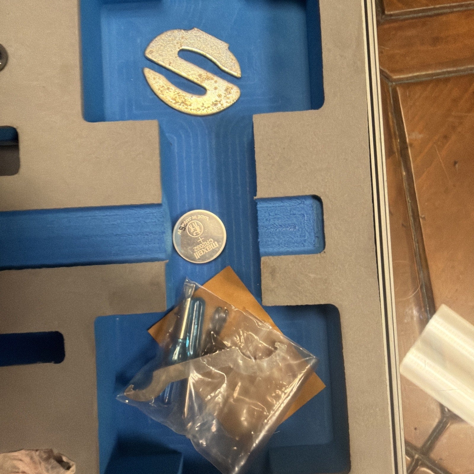 Blue foam insert with several small items including a round silver coin cell battery, a large metallic S-shaped piece, and a small plastic bag containing screws and metal parts, all placed inside a gray case on a wooden floor.