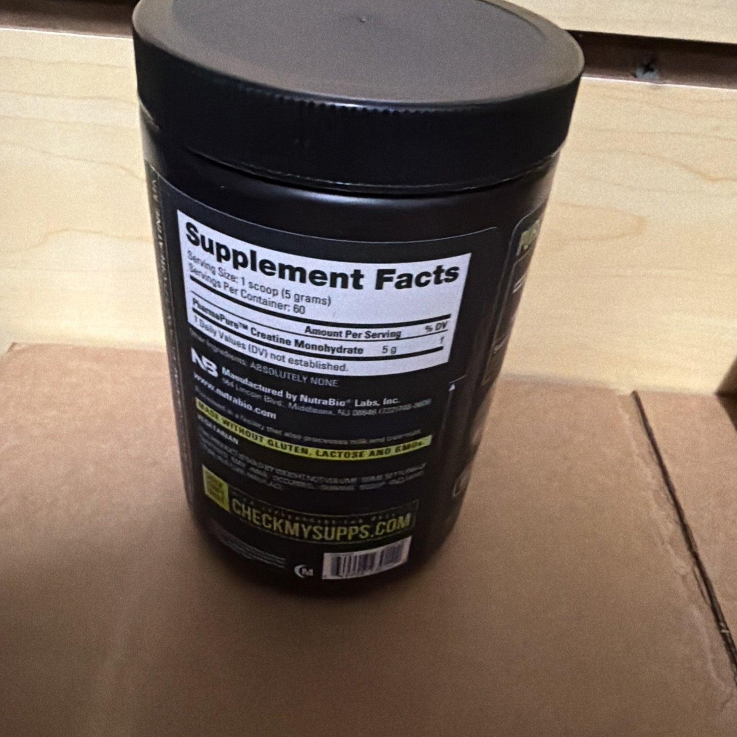Black plastic container with a close-up view of the supplement facts label showing creatine monohydrate content of 5 grams per scoop, placed on a cardboard surface.