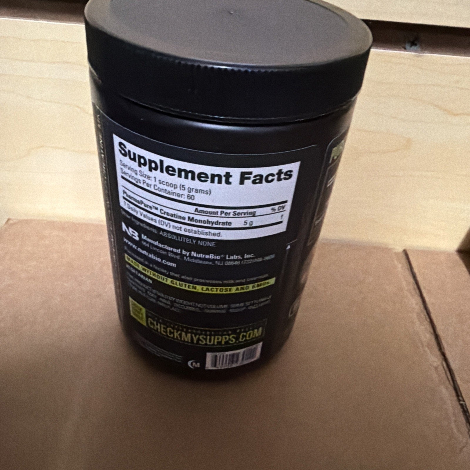 Black plastic container with a close-up view of the supplement facts label showing creatine monohydrate content of 5 grams per scoop, placed on a cardboard surface.