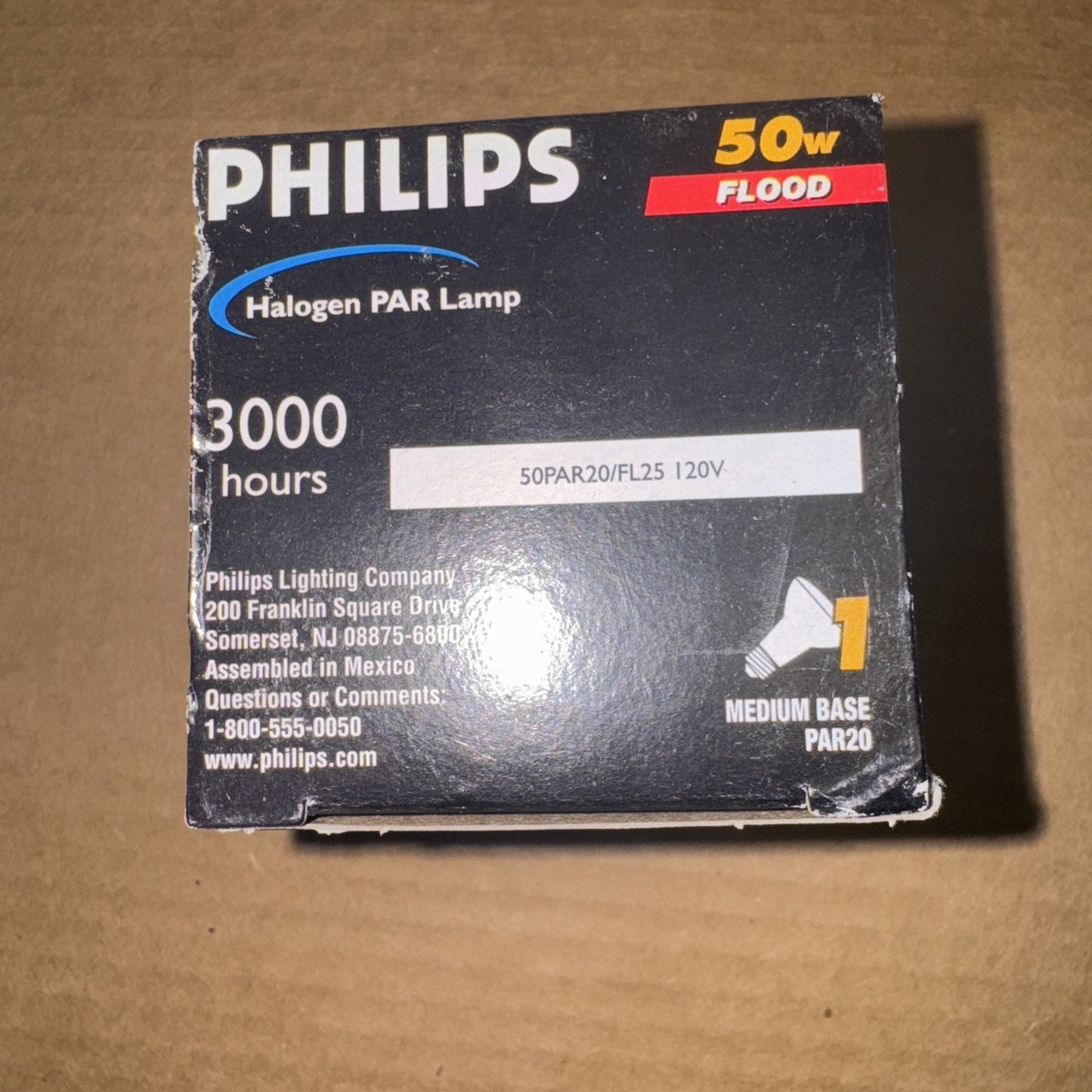 Black Philips box packaging for a 50 watt flood halogen PAR lamp with a medium base, rated for 3000 hours of use, showing text and contact information on a brown surface.