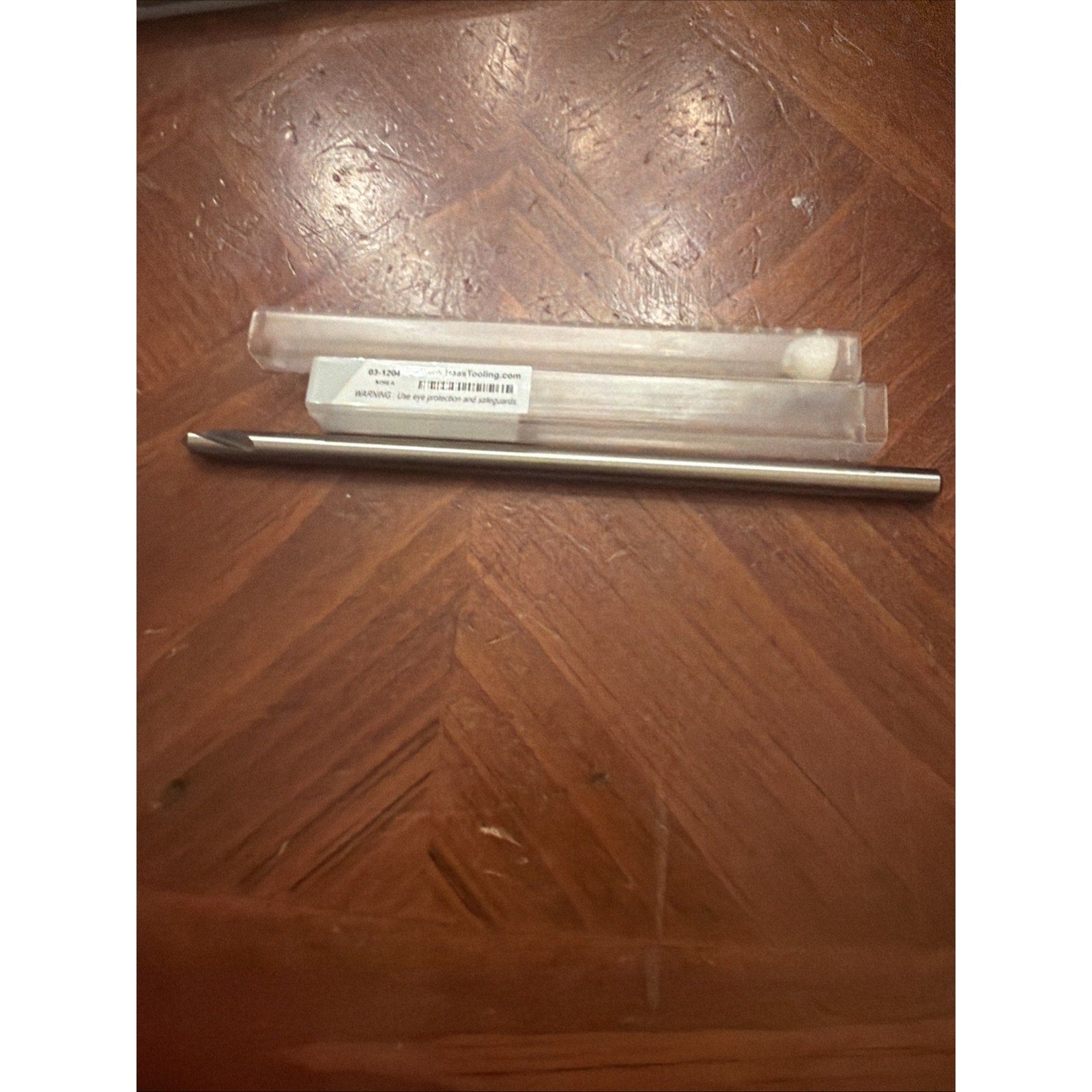 Metal rod tool with a pointed tip resting on a wooden surface near a clear plastic protective case with a white label containing printed text and barcode information.
