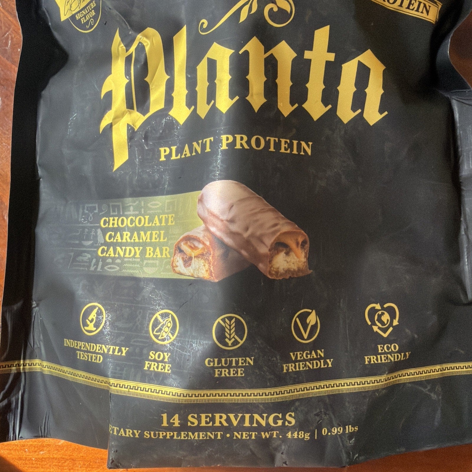 Black and gold packaging of Planta plant protein dietary supplement featuring chocolate caramel candy bar flavor, with icons indicating independently tested, soy free, gluten free, vegan friendly, and eco friendly, containing 14 servings and 448g net weight.