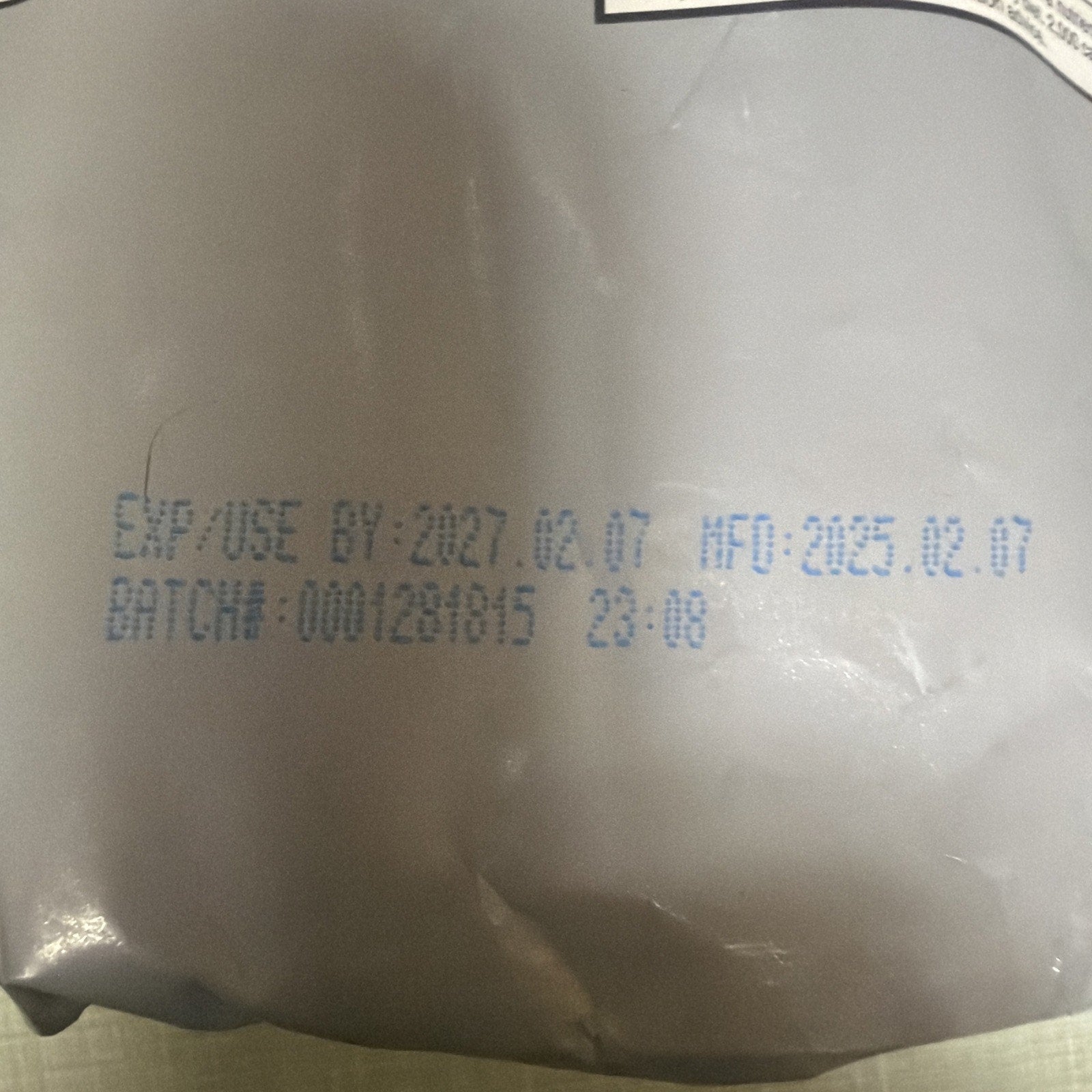 Close-up of packaging showing expiration date of February 7, 2027, manufacturing date of February 7, 2023, and batch number printed in blue ink on a silver surface.