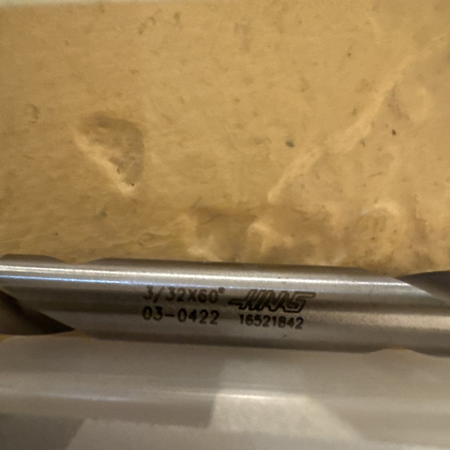 Close-up of a silver metal cutting tool engraved with "3/32X60," a logo, and numeric codes, resting against a textured beige surface and partially inside a white container.