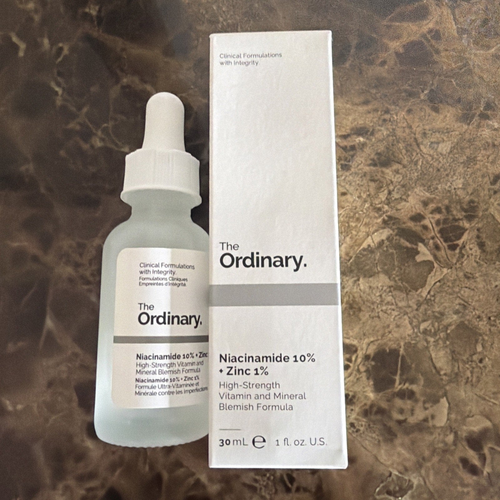 Frosted glass dropper bottle and white rectangular box of The Ordinary Niacinamide 10% + Zinc 1% serum with minimal black text on a brown marble surface.