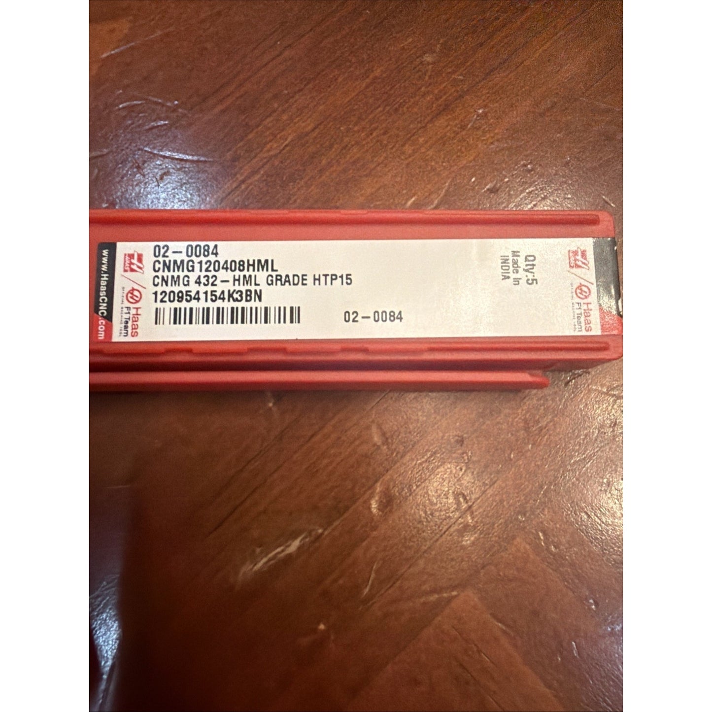 Red plastic case with a white label displaying product code "CNMG120408HML," grade "HTP15," barcode, and additional numeric identifiers, resting on a wooden surface.