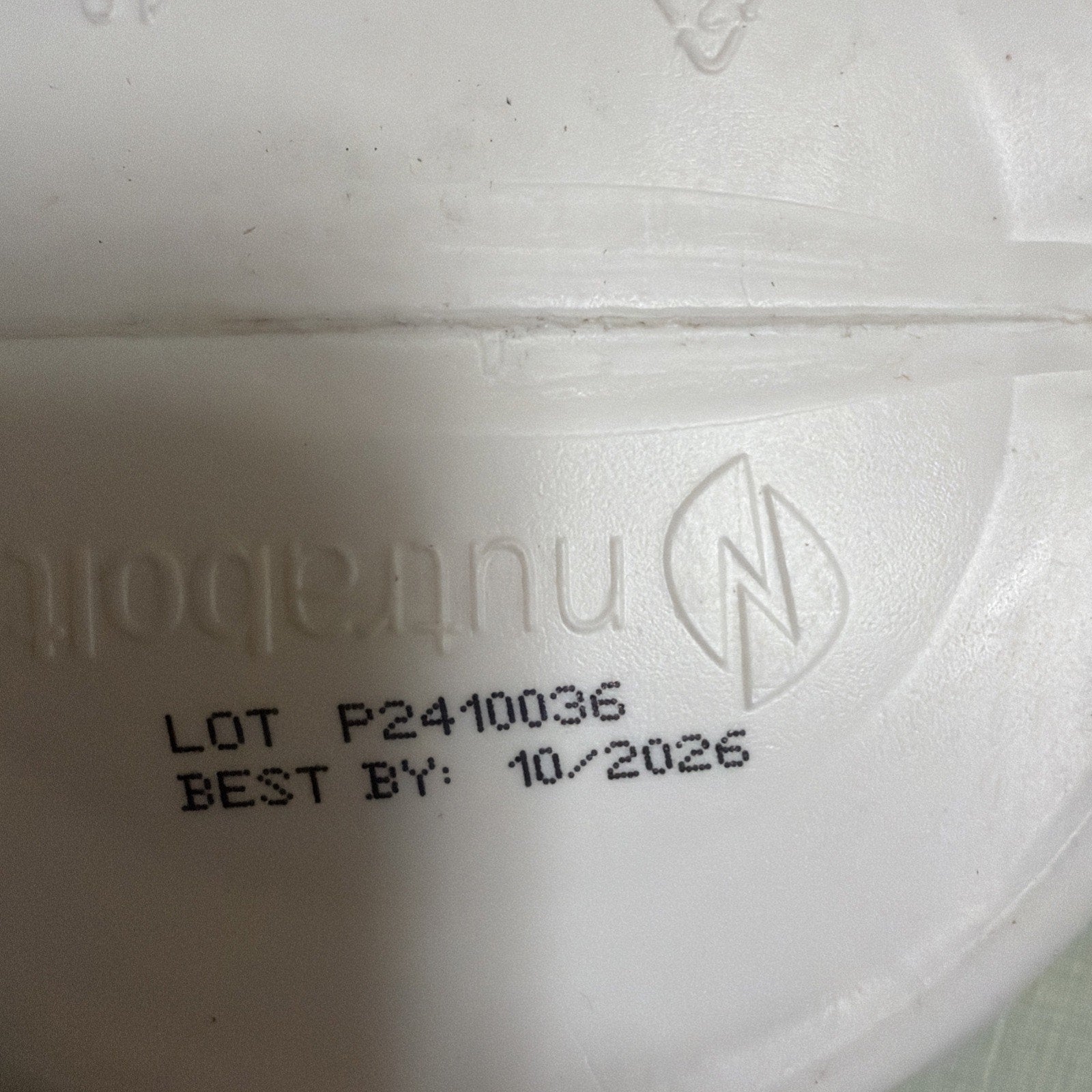 Close-up of a white plastic container lid showing printed lot number P2410036 and best by date 10/2026 on a textured surface with a faint embossed logo.